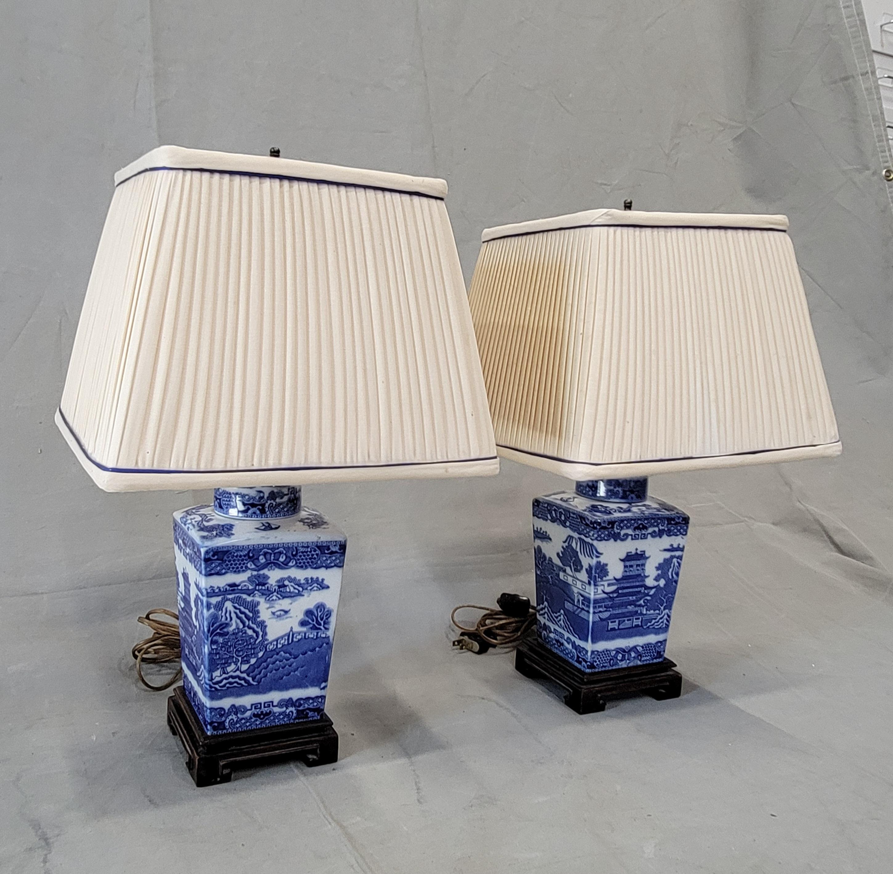 A charming pair of vintage 1950s blue willow lamps with original pleated ivory silk lampshades. The blue and white porcelain portion sits on top of carved wood bases. Wiring is original and works well. Pull chains and thumb switch both work. Made in