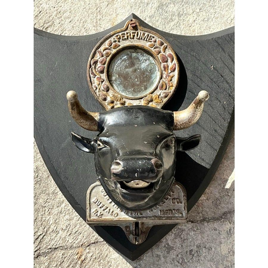 his captivating and rare vintage bull's head perfume dispenser hails from the early 1900s, a true statement piece blending artistry with functionality. Mounted on a shield-shaped wooden plaque, the intricately crafted bull's head features a central