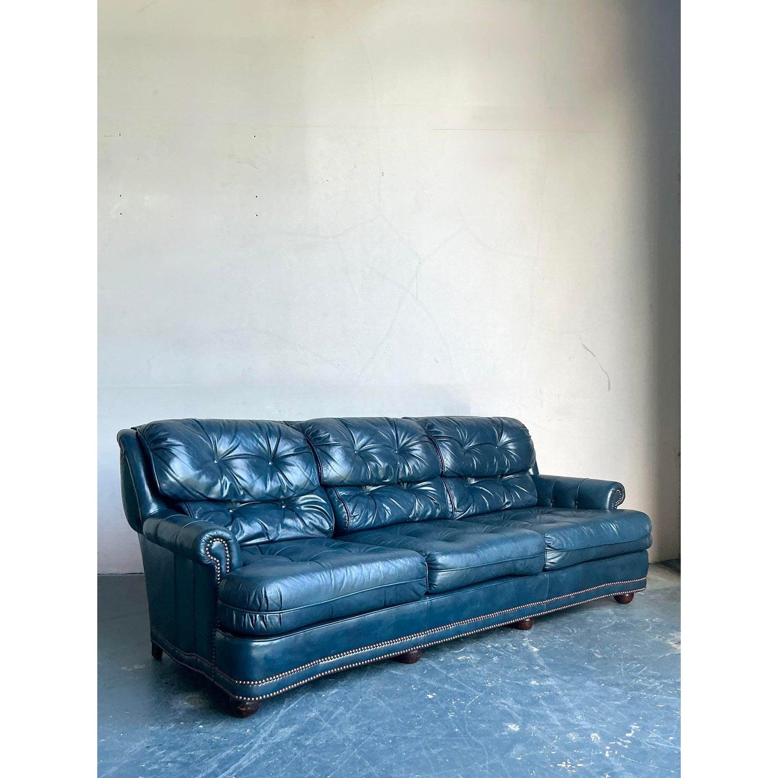 This imposing vintage sofa offers a luxurious and dramatic interpretation of the classic English Chesterfield, upholstered in a striking, rich petrol blue leather. The design features deep button tufting across the backrest and individual seat
