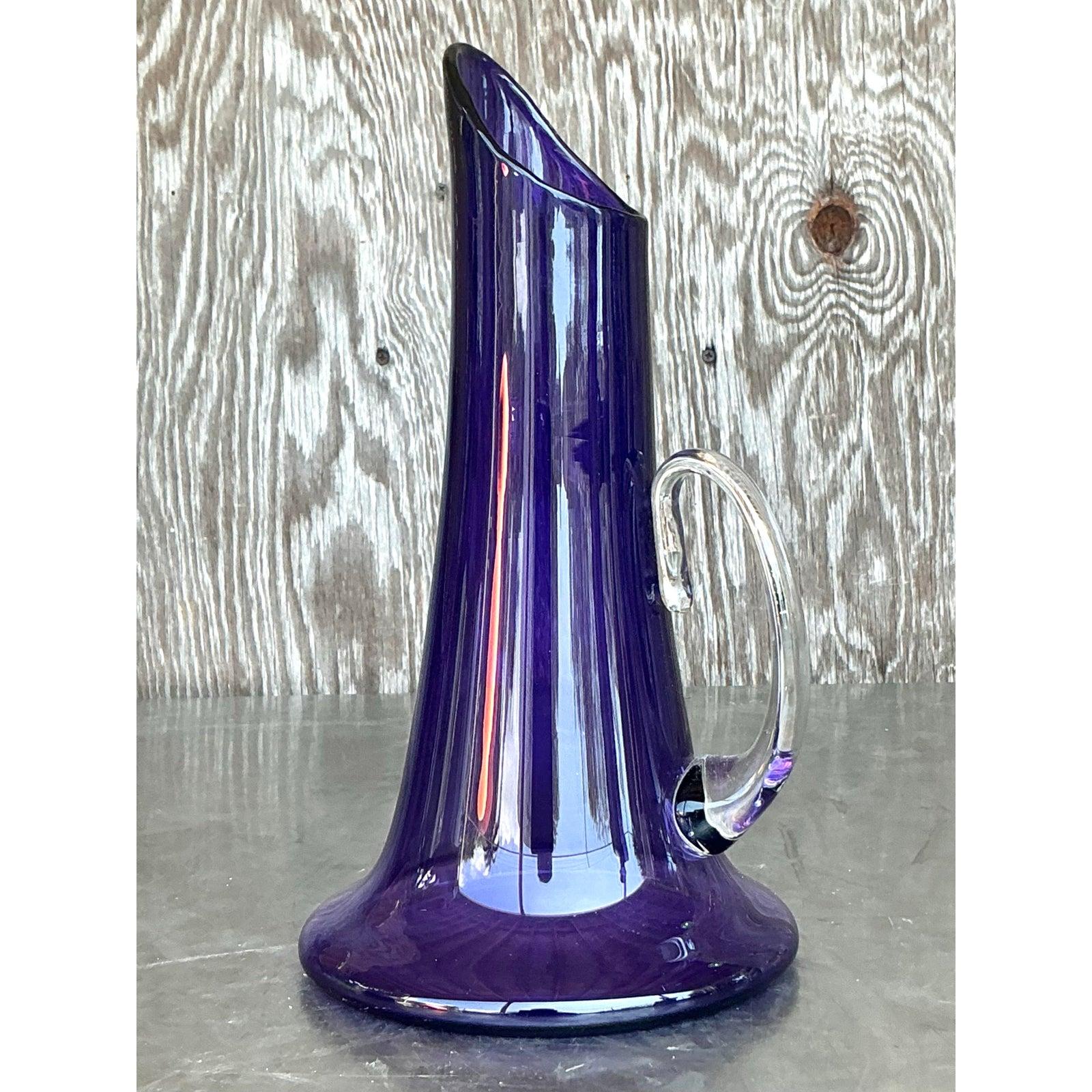 Bold in color and fluid in form, this Vintage Boho Art Glass Pitcher is a sculptural marvel that brings artistic flair to any space. With its rich amethyst hue and clear glass handle, the piece showcases expert craftsmanship and a dynamic