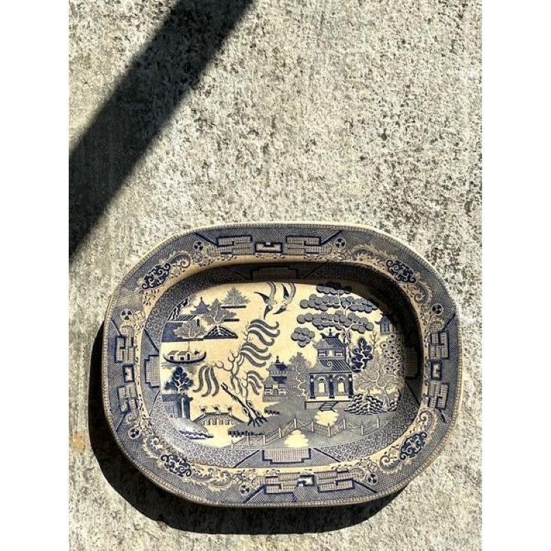 This stunning vintage Blue Willow ceramic platter showcases the timeless beauty of traditional transferware. Featuring an intricate blue and white chinoiserie landscape with pagodas, bridges, and willow trees, this piece tells a rich and storied