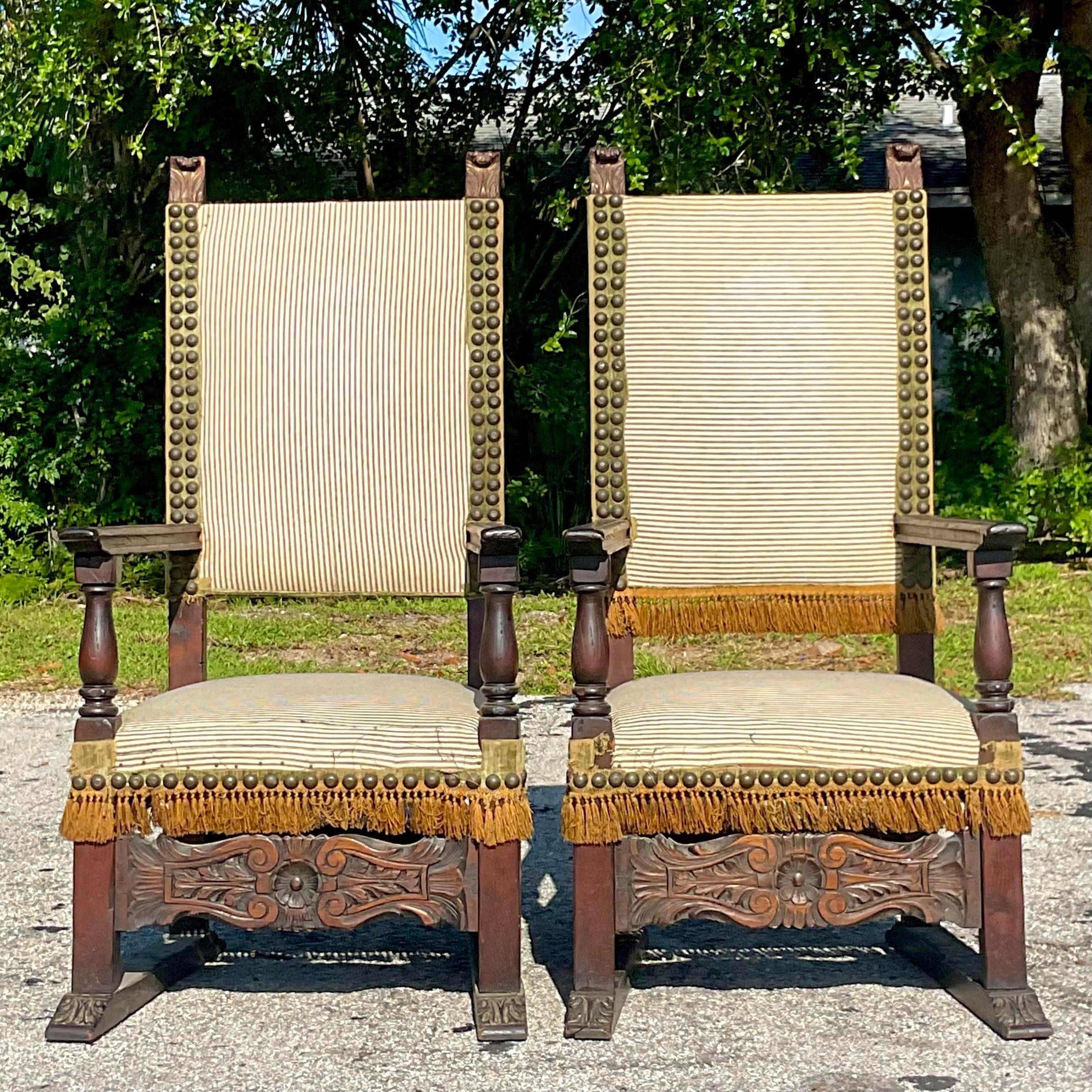 An extraordinary pair of vintage Boho Spanish high back chairs. Hand carved detail and a giant nailhead trim. A mattress ticking upholstery with a golden fringe. These chairs have it all. Acquired from a Palm Beach estate.