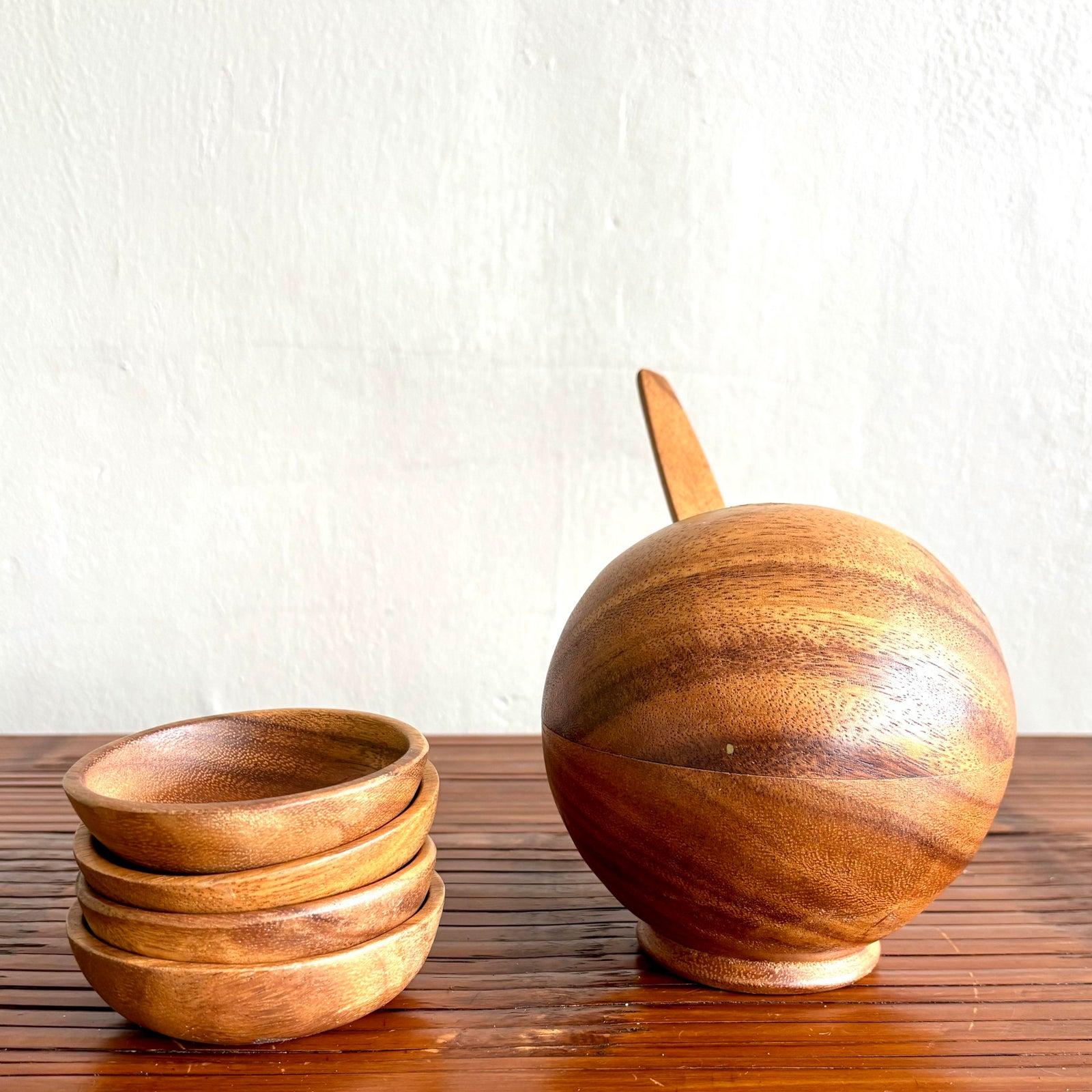 Dieses Set aus geschnitztem Holzgeschirr besteht aus einer runden Schale mit Deckel und einem passenden Servierlöffel sowie vier kleinen Schalen aus glattem, honigfarbenem Holz mit sichtbarer natürlicher Maserung. Das Design ist sowohl funktional