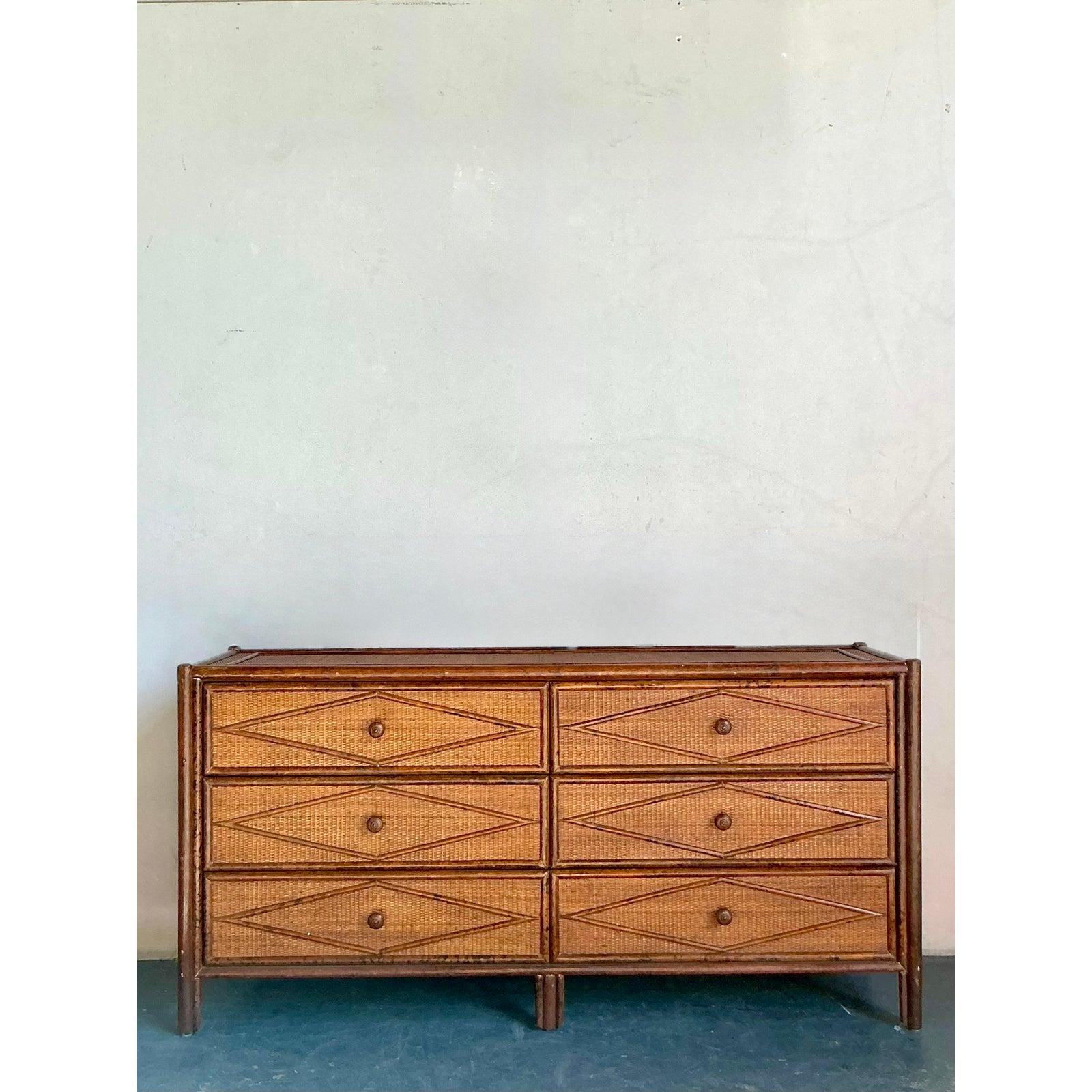 Ti presentiamo uno splendido comò vintage Boho Chic, magistralmente realizzato in bambù con finitura tartaruga e dotato di squisiti frontali dei cassetti in rattan con motivo a diamante. Questo imponente pezzo incarna una perfetta fusione di
