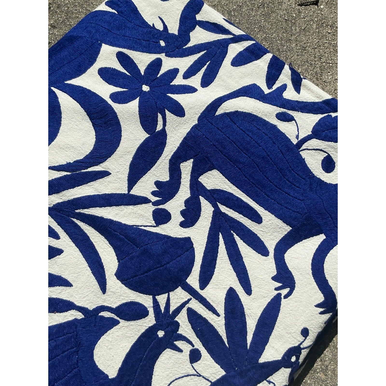 Infuse your dining space with artisanal charm with this stunning vintage embroidered tablecloth. Showcasing an intricate hand-stitched design in a bold cobalt blue on crisp white fabric, this textile reflects the artistry and craftsmanship of