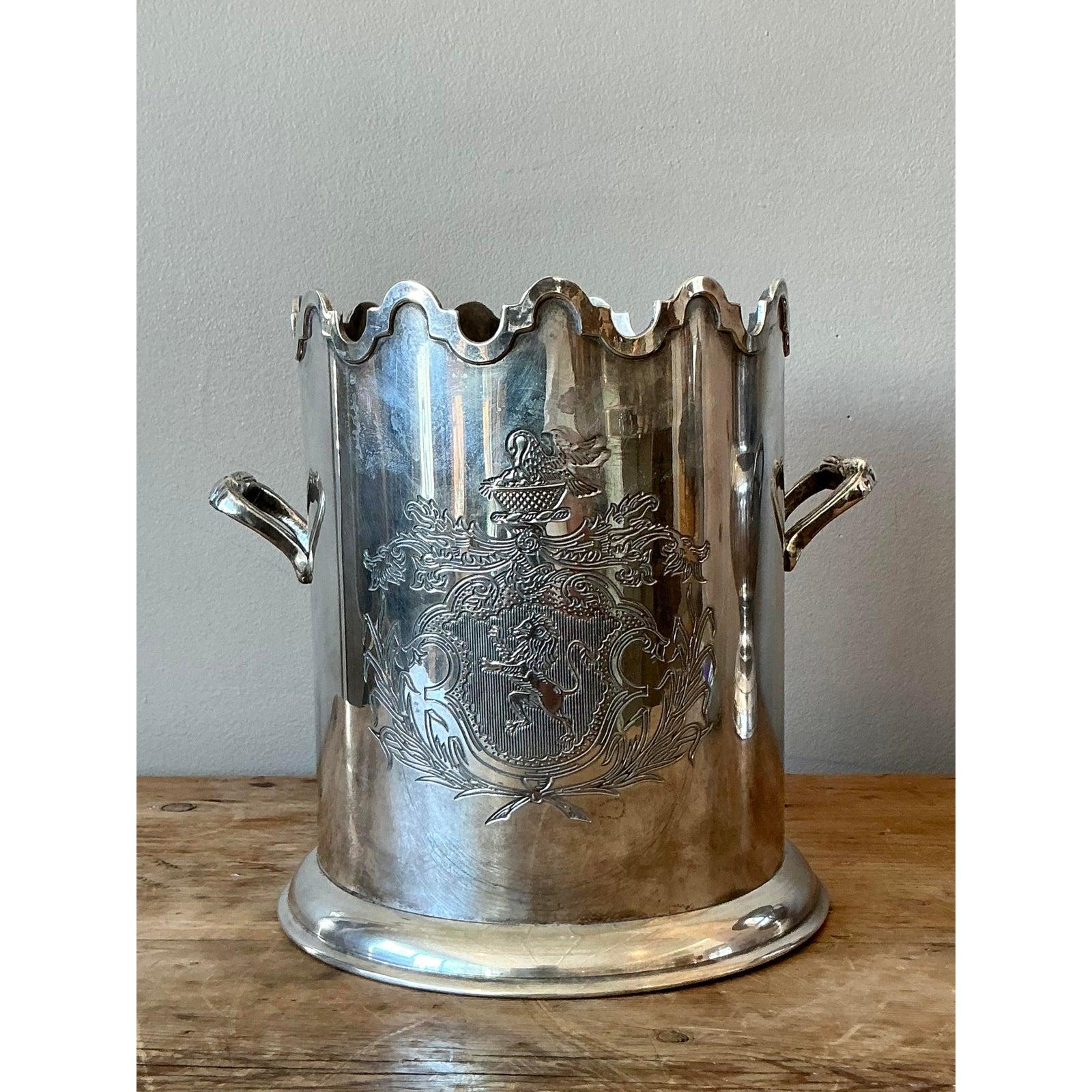 This stately and elegant vintage champagne bucket is a magnificent example of timeless silverplate craftsmanship. The piece features a polished finish, an ornate scalloped rim, and two beautifully detailed scrolled handles. The centerpiece is a