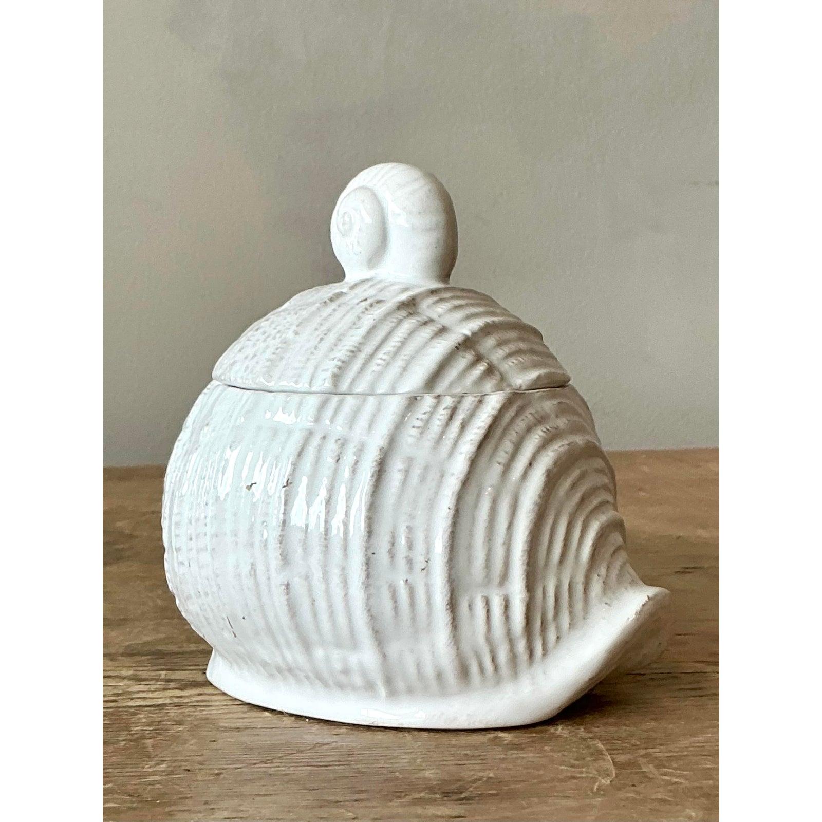 Insufflez à votre rituel matinal la beauté sereine de la côte grâce à ce charmant pot de miel vintage en céramique. Sculpté sous la forme d'une conque ou d'un turban en spirale, le récipient présente une surface tactile et nervurée, animée par une