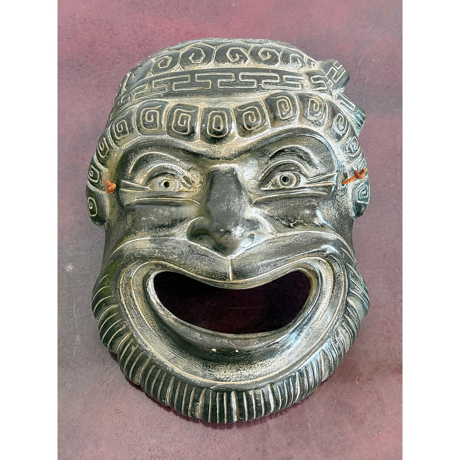 Una maschera vintage audace ed espressiva ispirata al teatro greco classico, caratterizzata da una bocca spalancata, un sorriso esagerato e dettagli stilizzati e vorticosi sui capelli e sulle sopracciglia. La patina invecchiata e i dettagli