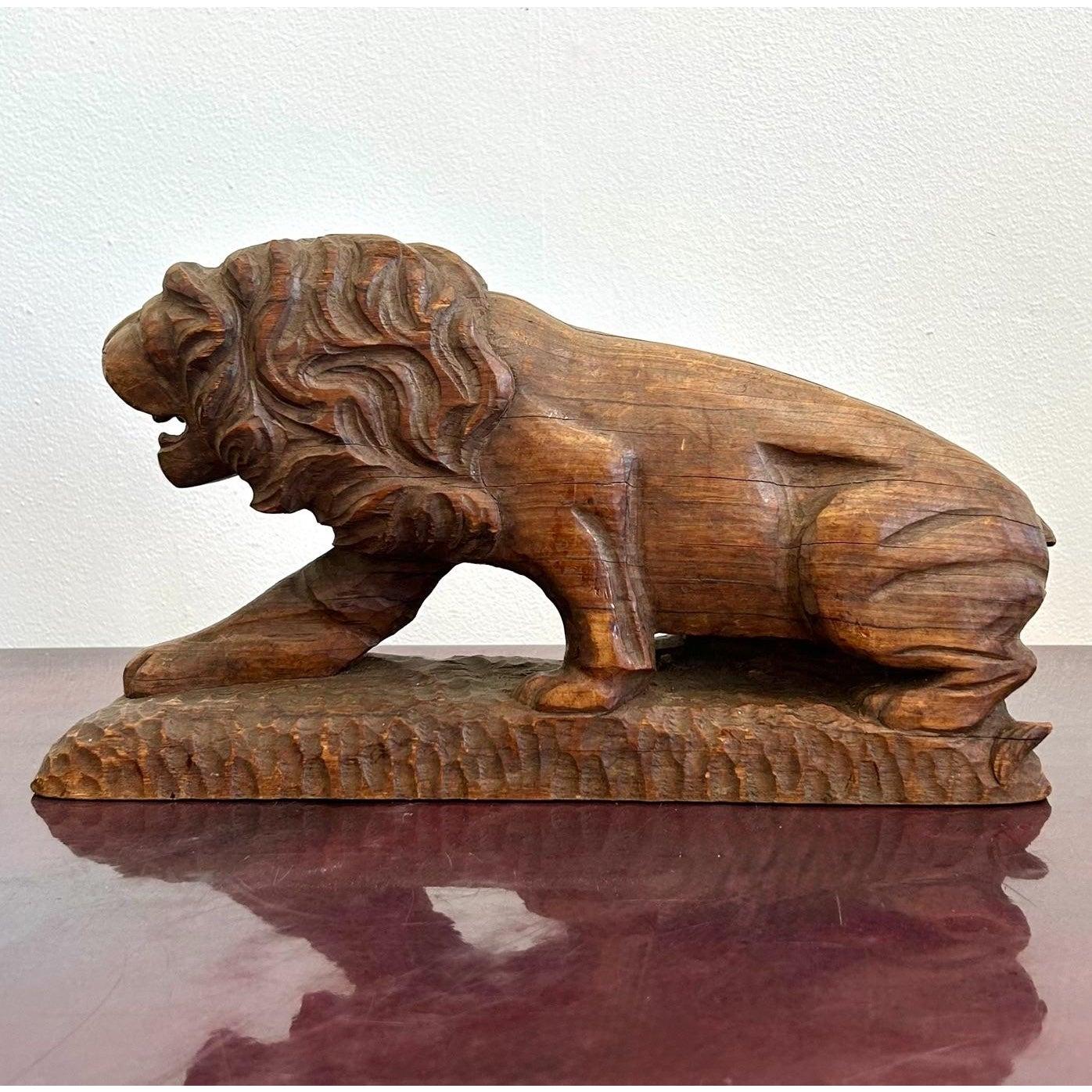 Questa scultura in legno intagliata a mano raffigura un leone in posizione accovacciata e possente, con una zampa protesa in avanti e la testa leggermente chinata, come se fosse nel pieno del ruggito o in un momento di tensione. La folta e fluente