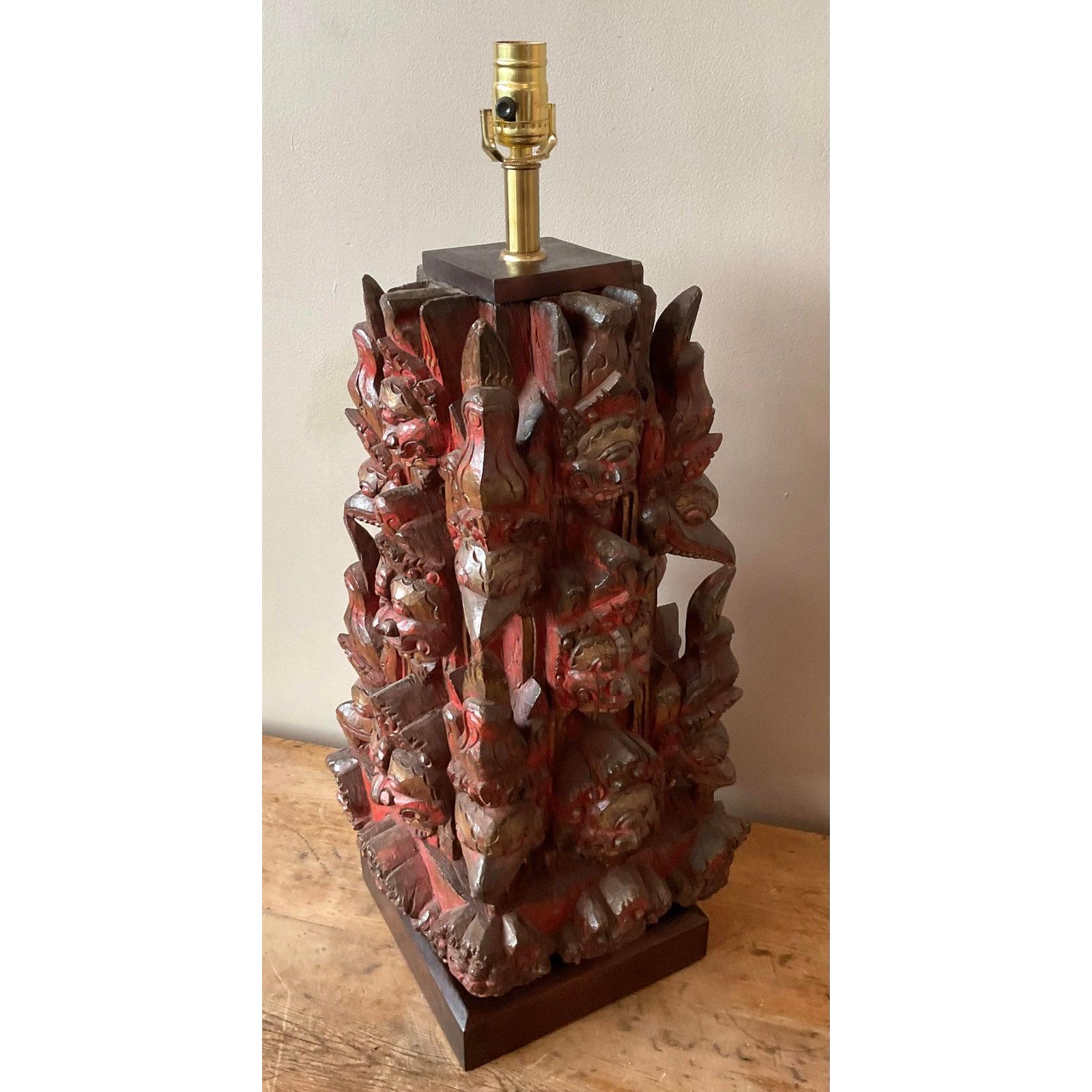 This powerful and intricate vintage table lamp is a masterpiece of hand-carved folk art. Featuring a complex, multi-faced temple mask design, the lamp base is a captivating sculpture crafted from wood and adorned with red, gold, and black pigments.