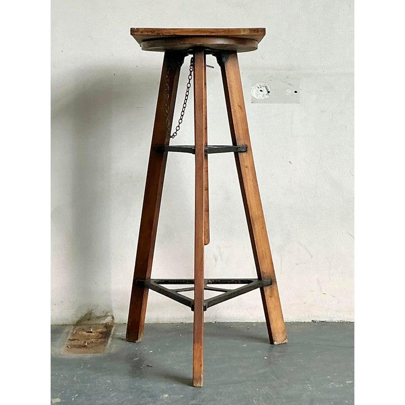 Discover the captivating blend of artistry and industrial design with this unique vintage adjustable display pedestal. The pedestal features a robust wooden tripod base, accented with dark metal stretchers and an adjustable mechanism, evoking a