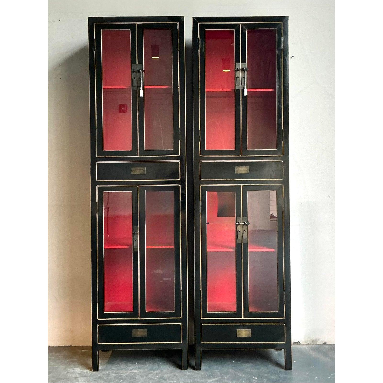 Presenting a stunning pair of vintage Boho Chic display cabinets, meticulously crafted with a captivating black lacquered finish. These tall, elegant cabinets exude an exotic charm, highlighted by delicate gold-painted accents that trace their