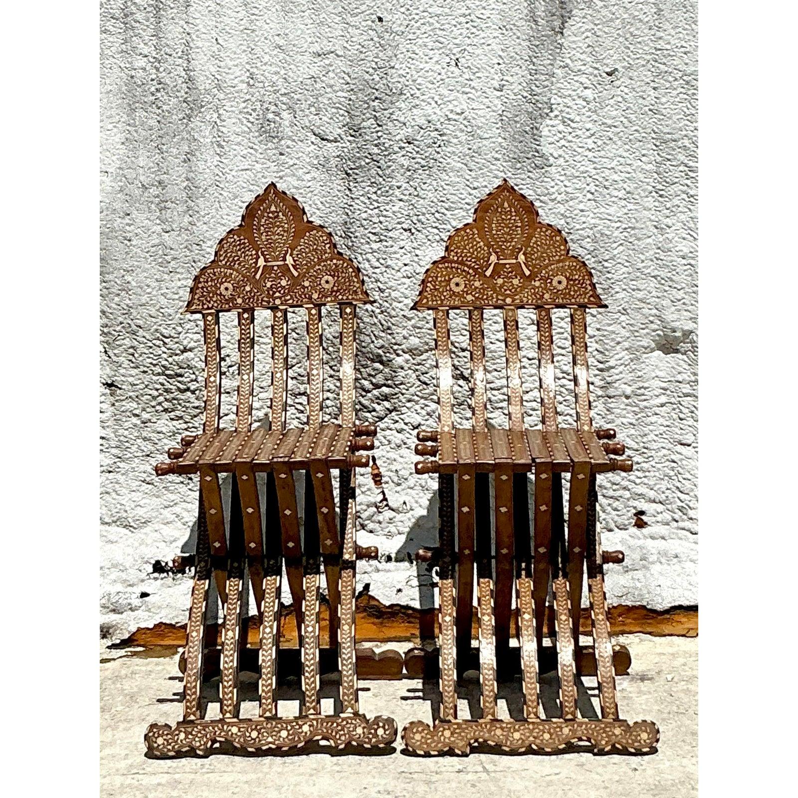 Unfold a world of exotic charm with this exquisite pair of Vintage Boho Moorish Inlay Folding Chairs. Each chair is a testament to extraordinary craftsmanship, featuring intricate mother-of-pearl inlay and delicate carved details that tell a story