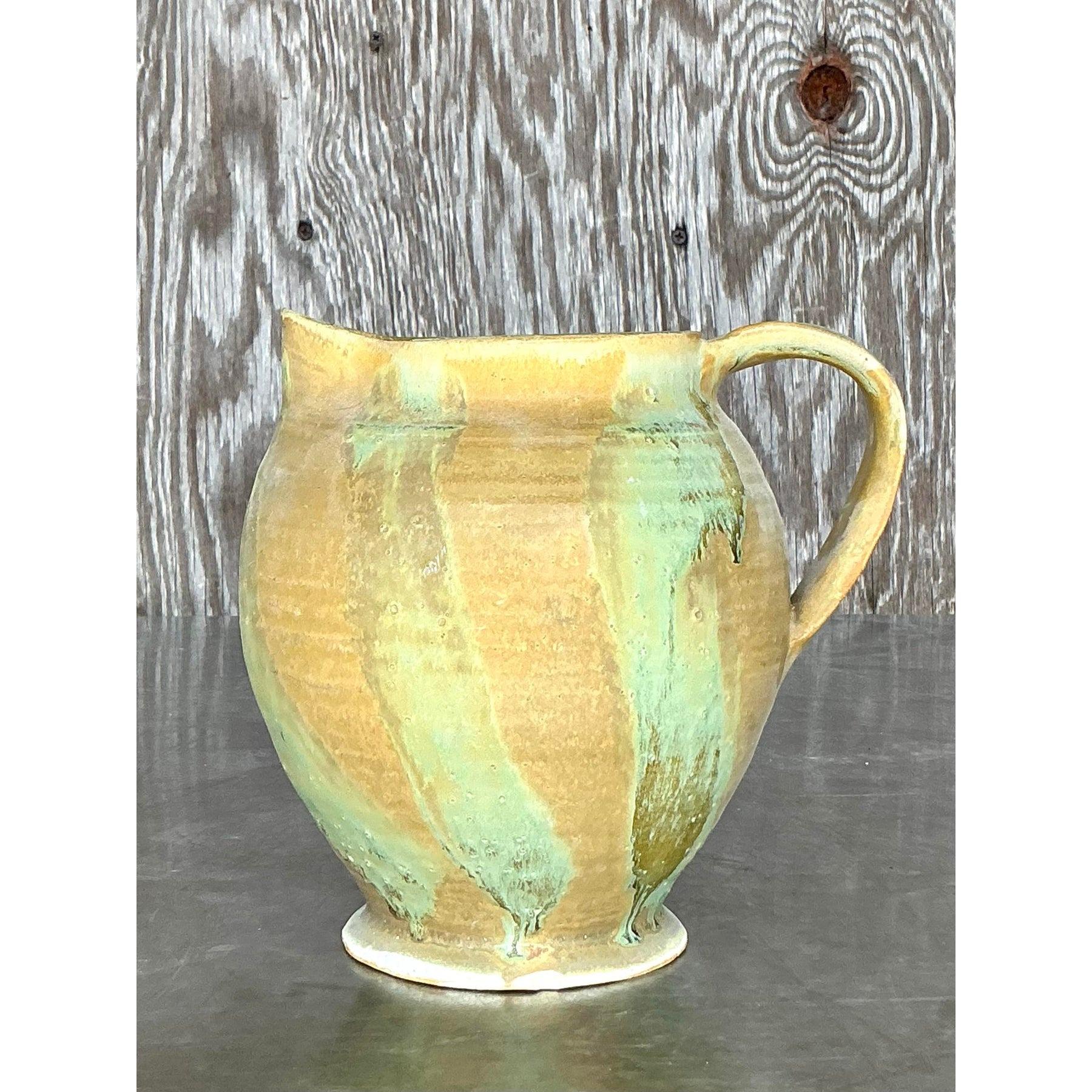 Add a touch of handcrafted artistry to your space with this vintage boho original studio pottery pitcher. This beautifully thrown ceramic piece features an earthy glaze blending soft greens, sandy yellows, and subtle blue undertones, creating a