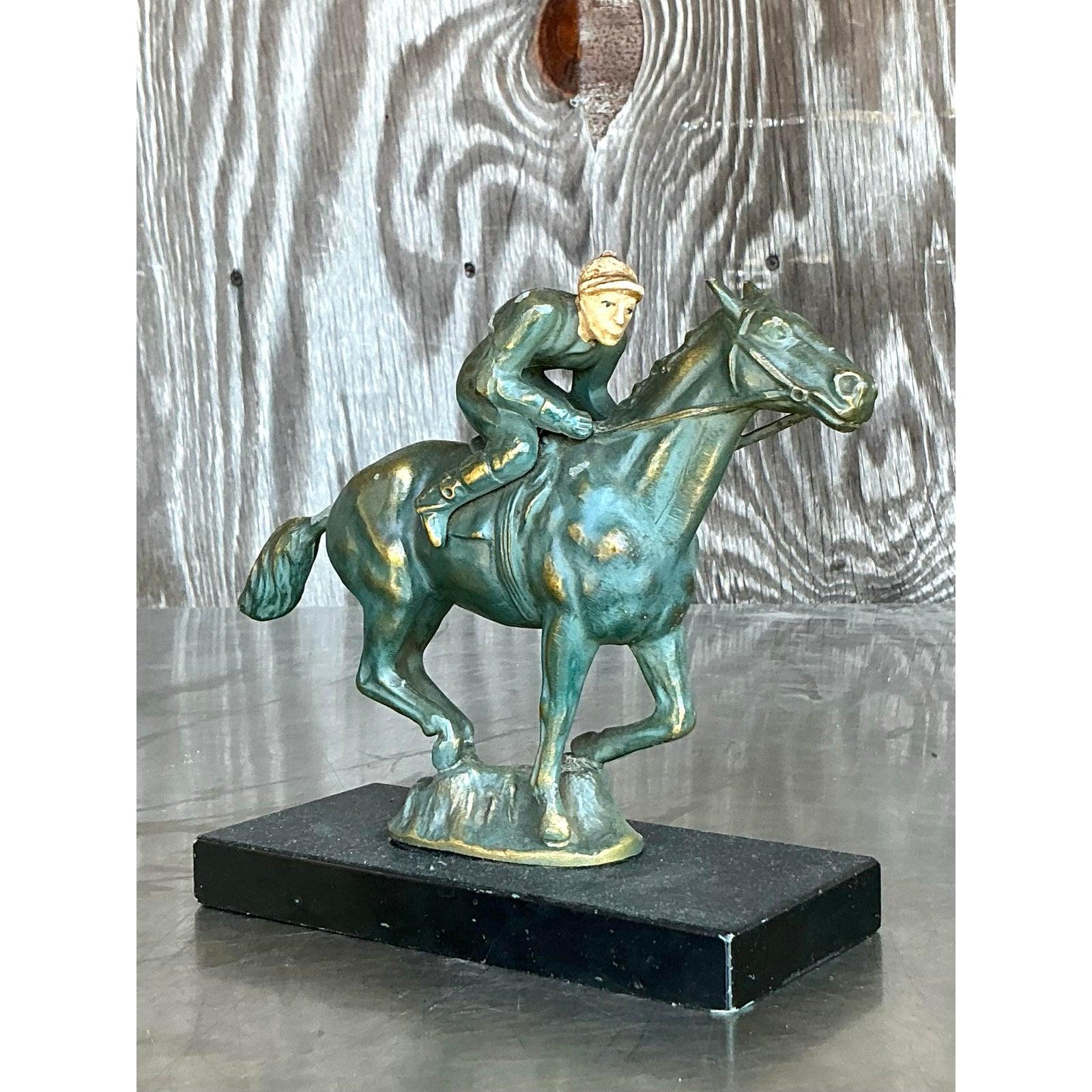 Cattura lo spirito della velocità e della grazia con questa scultura vintage in ottone Boho che raffigura un fantino in piena corsa in cima a un cavallo al galoppo. Questo pezzo in ottone patinato è pieno di movimento ed energia, con dettagli