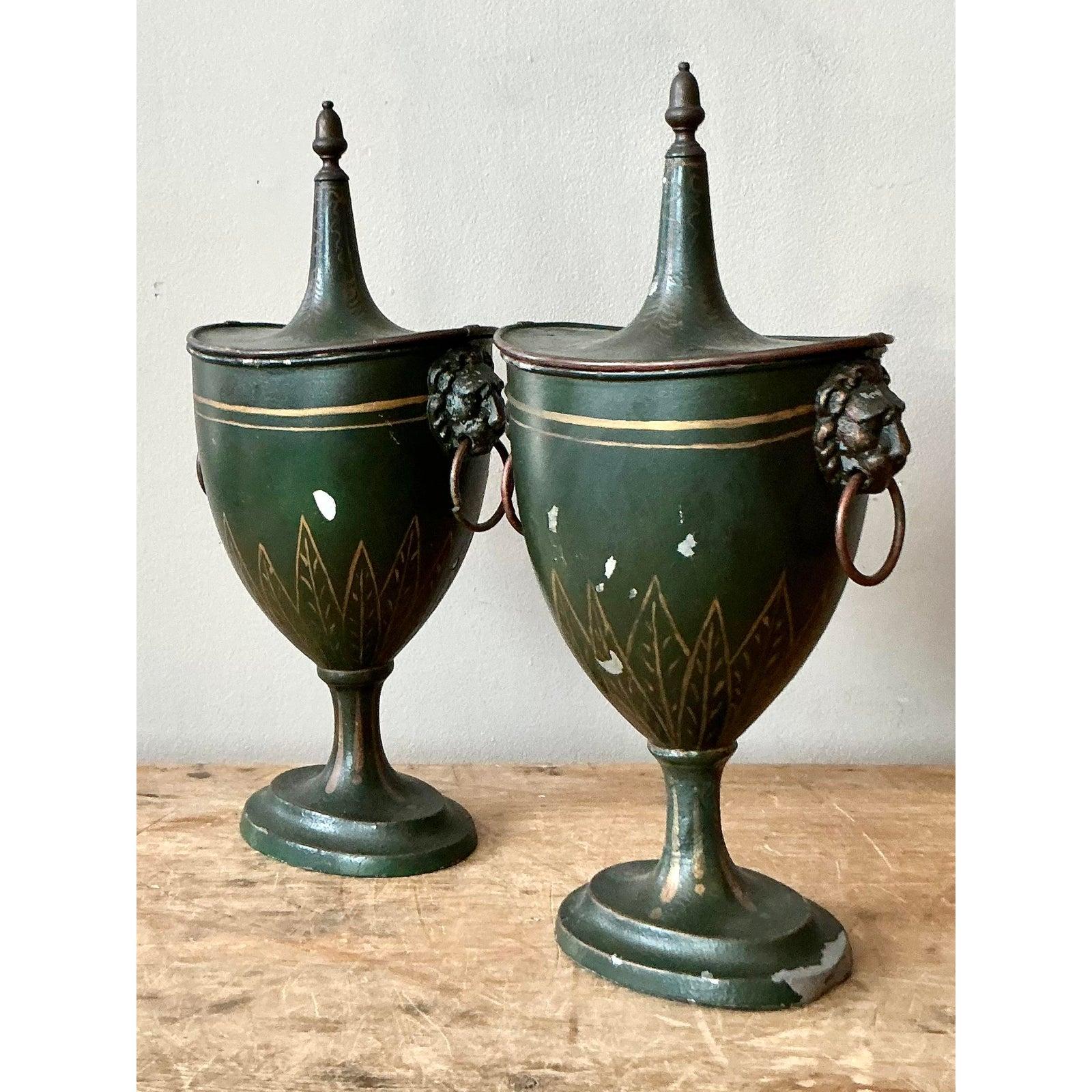 Questa bella coppia di urne decorative cattura la maestosa eleganza del design neoclassico, utilizzando il metallo verniciato (Tole Peinte) per ottenere un'estetica sofisticata e invecchiata. Ogni urna è caratterizzata da una classica Silhouette con