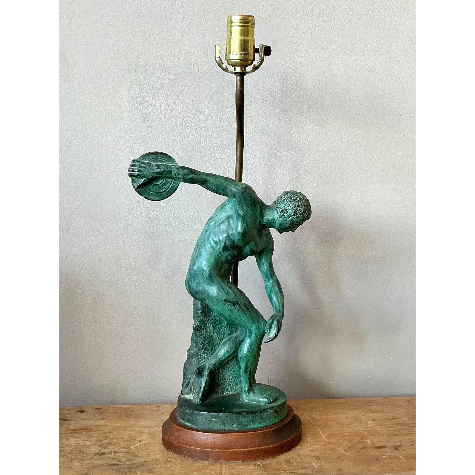 Esta distinguida lámpara de sobremesa rinde homenaje a la antigüedad clásica, con una llamativa base figurada inspirada en el célebre Discóbolo (lanzador de discos) de Mirón. La escultura, fundida en metal, está tratada con una rica pátina