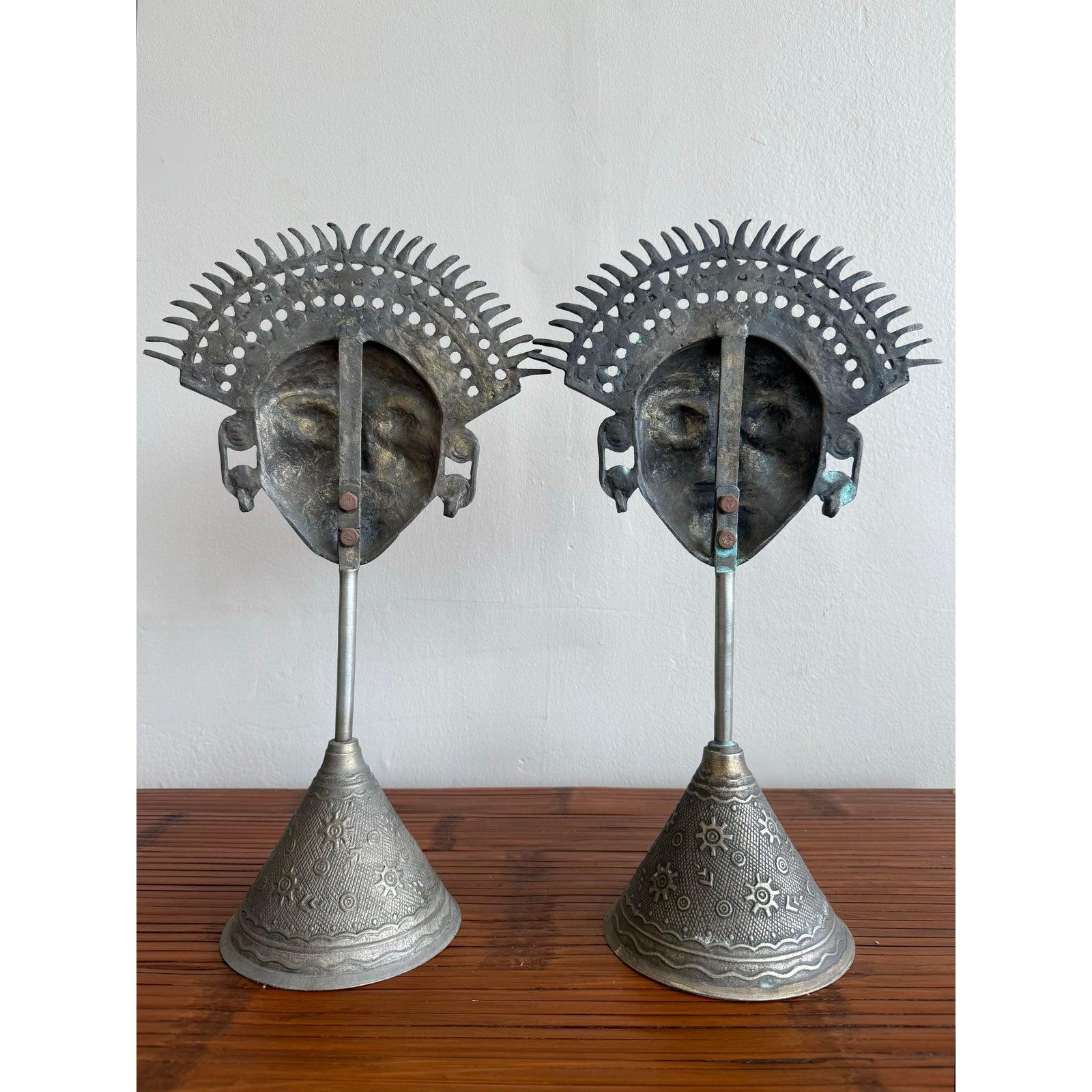 Queste teste scultoree in metallo emanano carattere e presenza culturale con i loro intricati copricapi, i volti espressivi e le basi a cono finemente modellate. L'accoppiamento simmetrico suggerisce la dualità o l'equilibrio, aggiungendo profondità