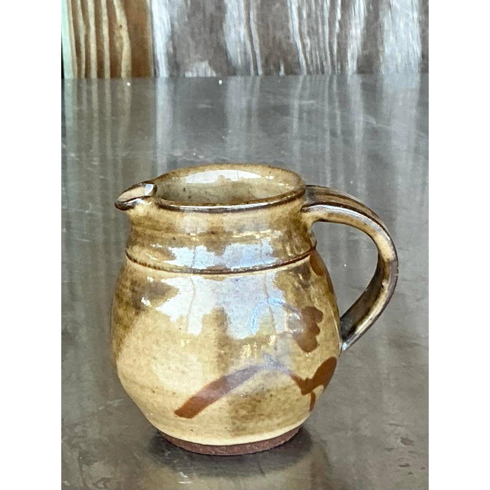 Add a touch of handcrafted charm to your daily rituals with this delightful vintage studio pottery pitcher. Its endearing, rounded form, complete with a comfortable handle and neat spout, is adorned with a warm, earthy glaze and free-flowing