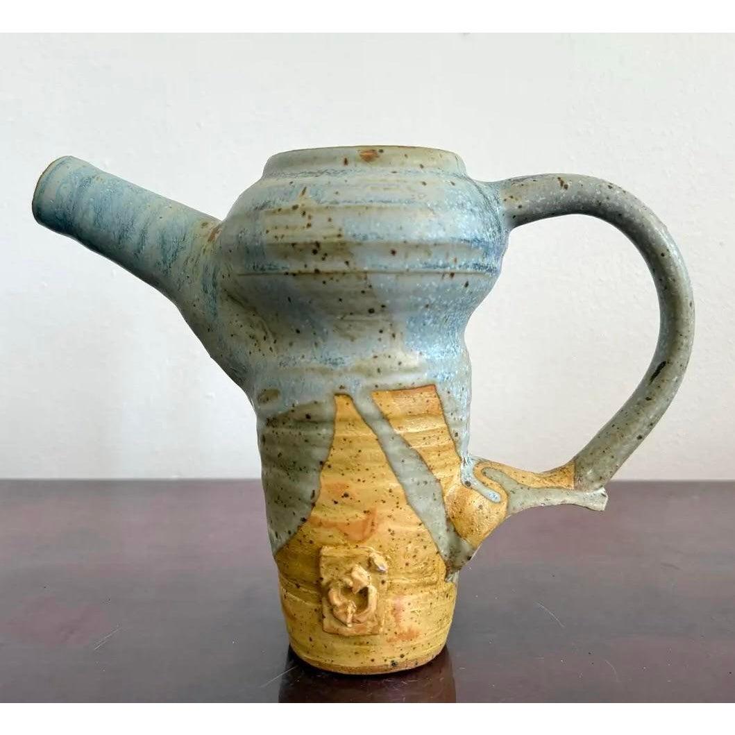 Esta caprichosa jarra de cerámica combina la funcionalidad con el estilo escultórico, y su caño y asa exagerados le confieren una postura juguetona, casi animada. El vidriado pasa de un suave azul celeste a cálidos tonos ocres, con goteos y motas
