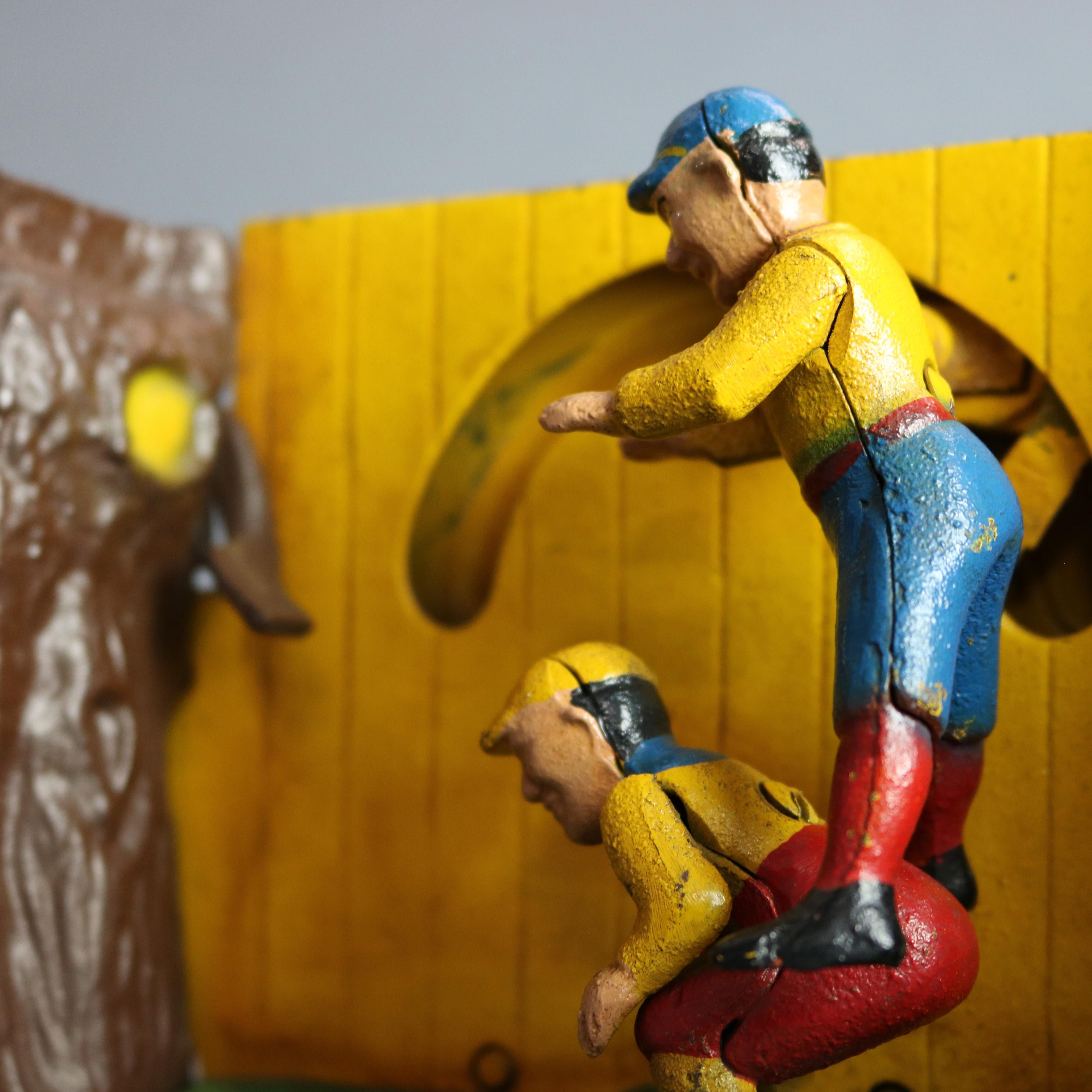 A vintage Book of Knowledge Leap Frog mechanical bank offers polychrome cast construction with boys playing leap frog outside by fence and tree with embossed title, underside reads 