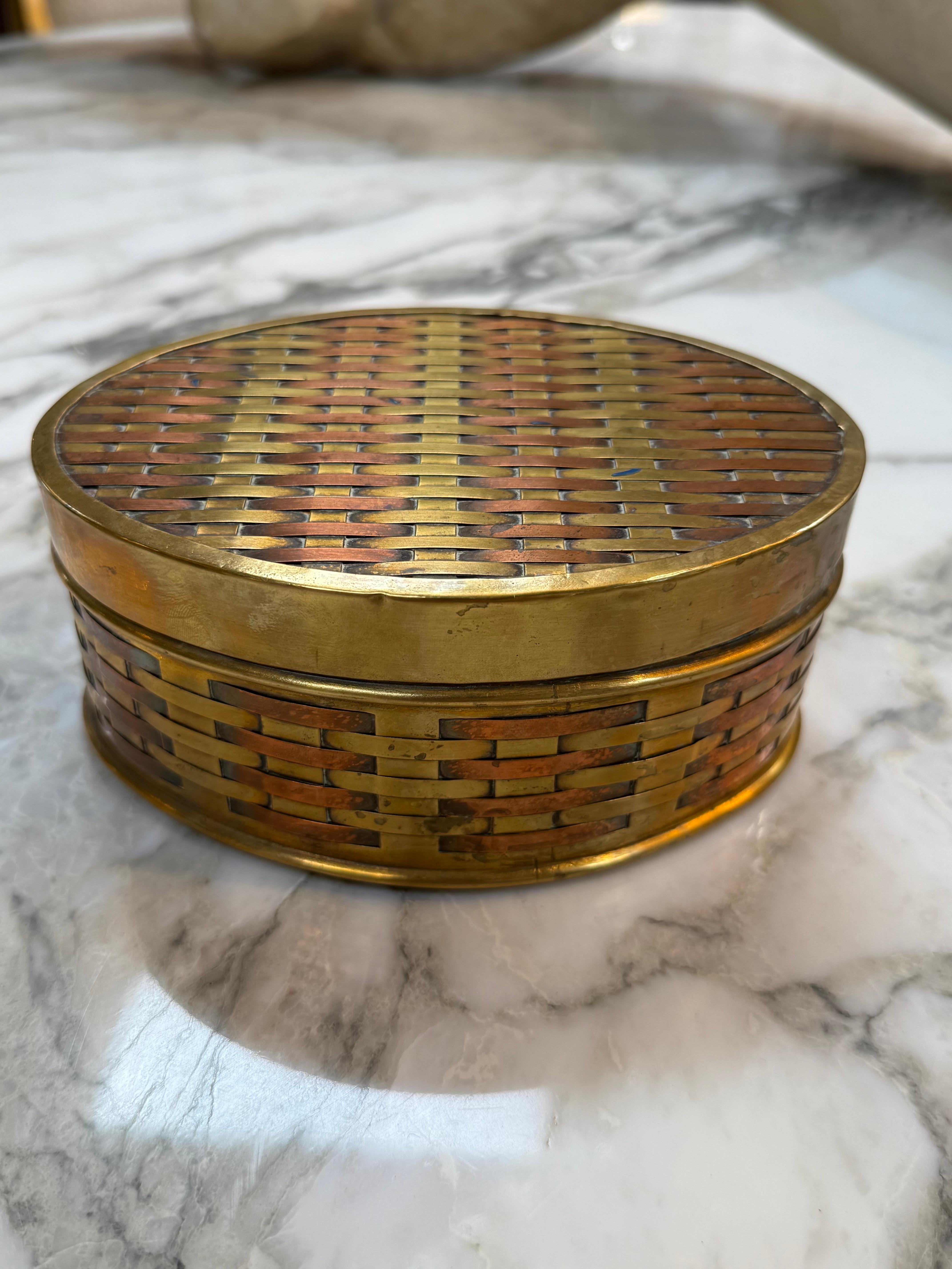 Questa scatola vintage rotonda presenta un intricato motivo intrecciato realizzato con strisce di ottone e rame alternate, creando un ricco gioco di calde tonalità metalliche. La texture simile a un cesto avvolge il corpo e il coperchio, dando al