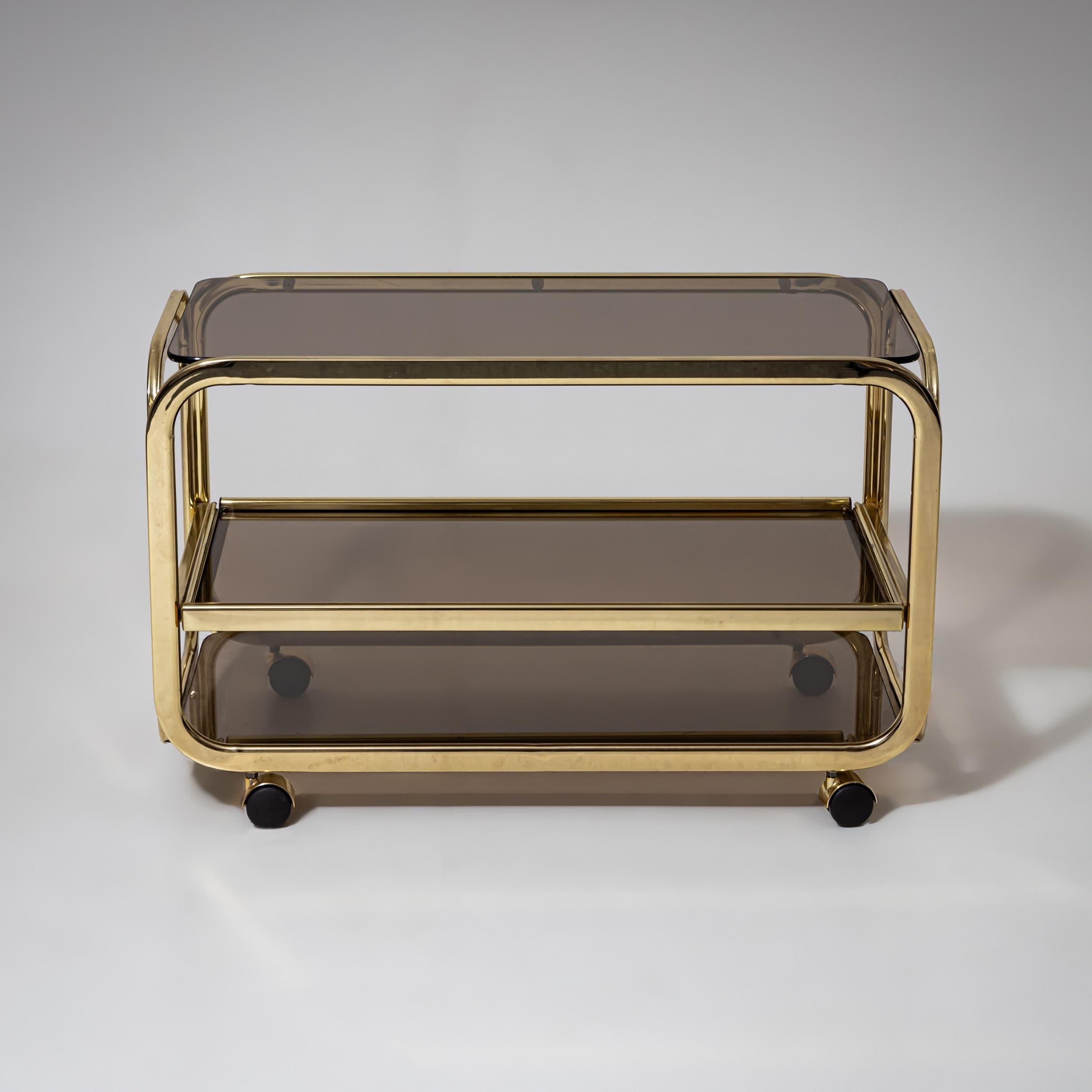 Stilvoller Barwagen aus den 1980er Jahren mit einem polierten Messingrahmen und drei Ablagen aus Rauchglas. Die abgerundete Silhouette verleiht ihm ein Gefühl von raffinierter Leichtigkeit, während die Rollen für einfache Mobilität sorgen. Ein