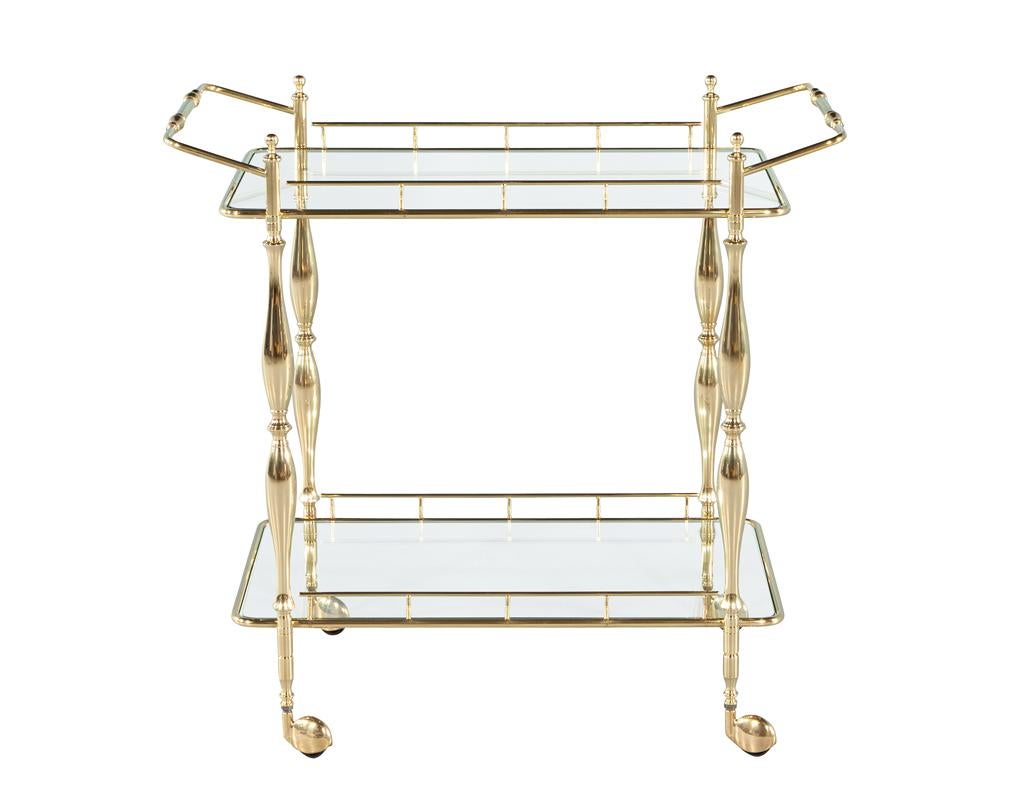 Transport yourself back to the glamorous 1980’s with this stunning vintage brass bar cart. Crafted in the USA, this piece embodies classic traditional style, featuring polished brass framing that gleams beautifully. Two sleek glass shelves provide