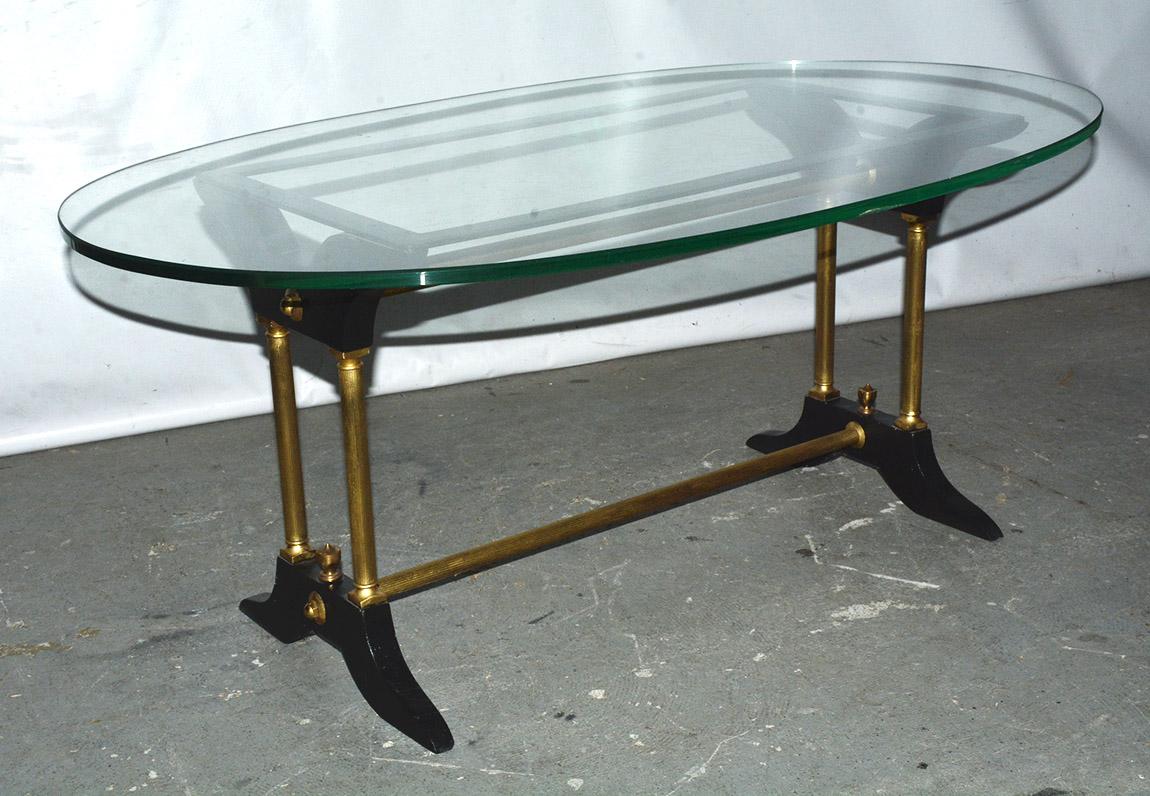 The vintage coffee table has a base made of brass fluted tubing that connects black wood feet and braces, the latter of which holds a rectangular black iron frame upon which the oval glass top rests - contemporary with classical details. Table base