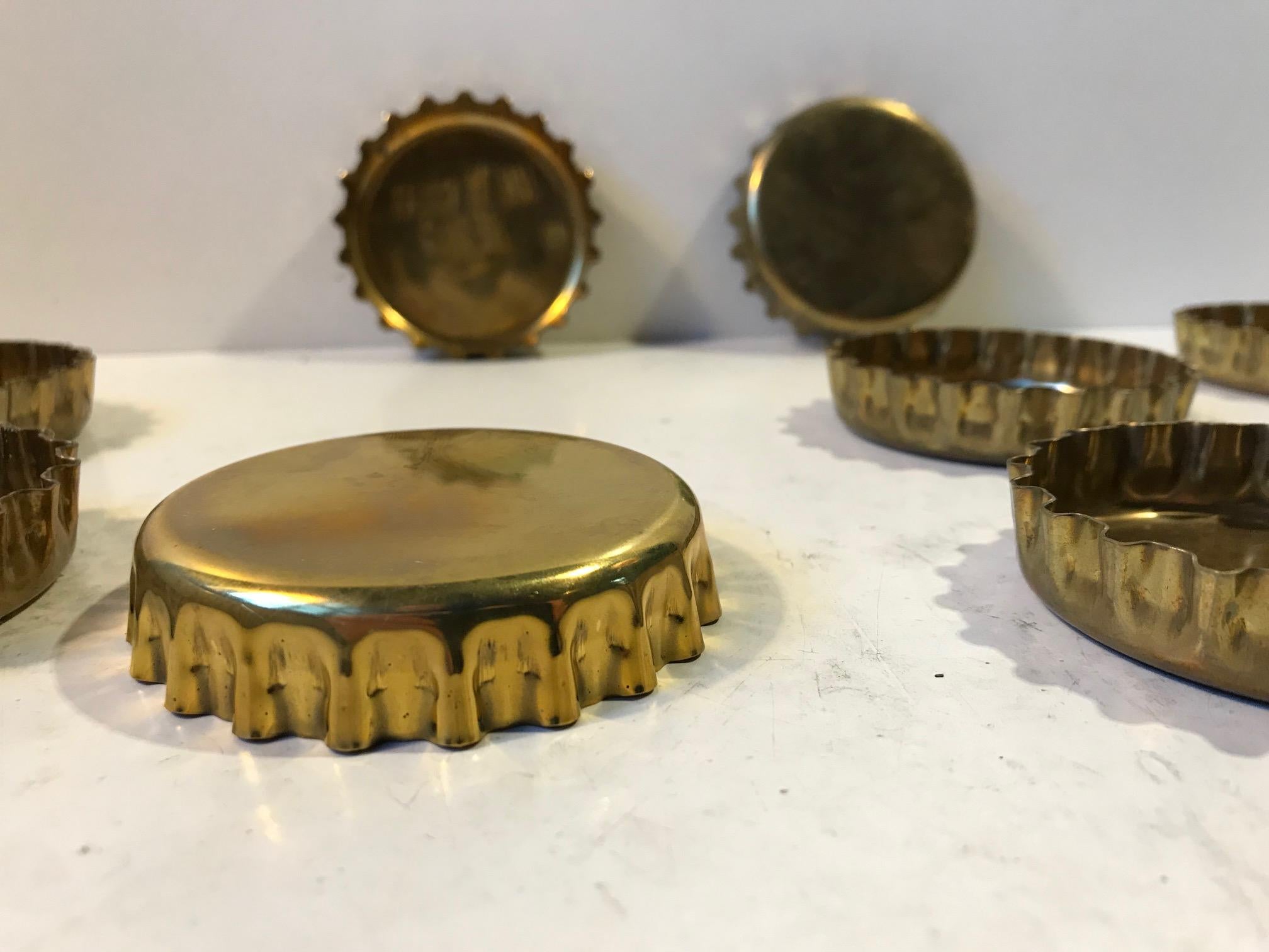 Set of 8 drink coasters in brass that are shaped as bottle caps. They are designed and manufactured by Georg Jensen in Denmark during the early 1980s.