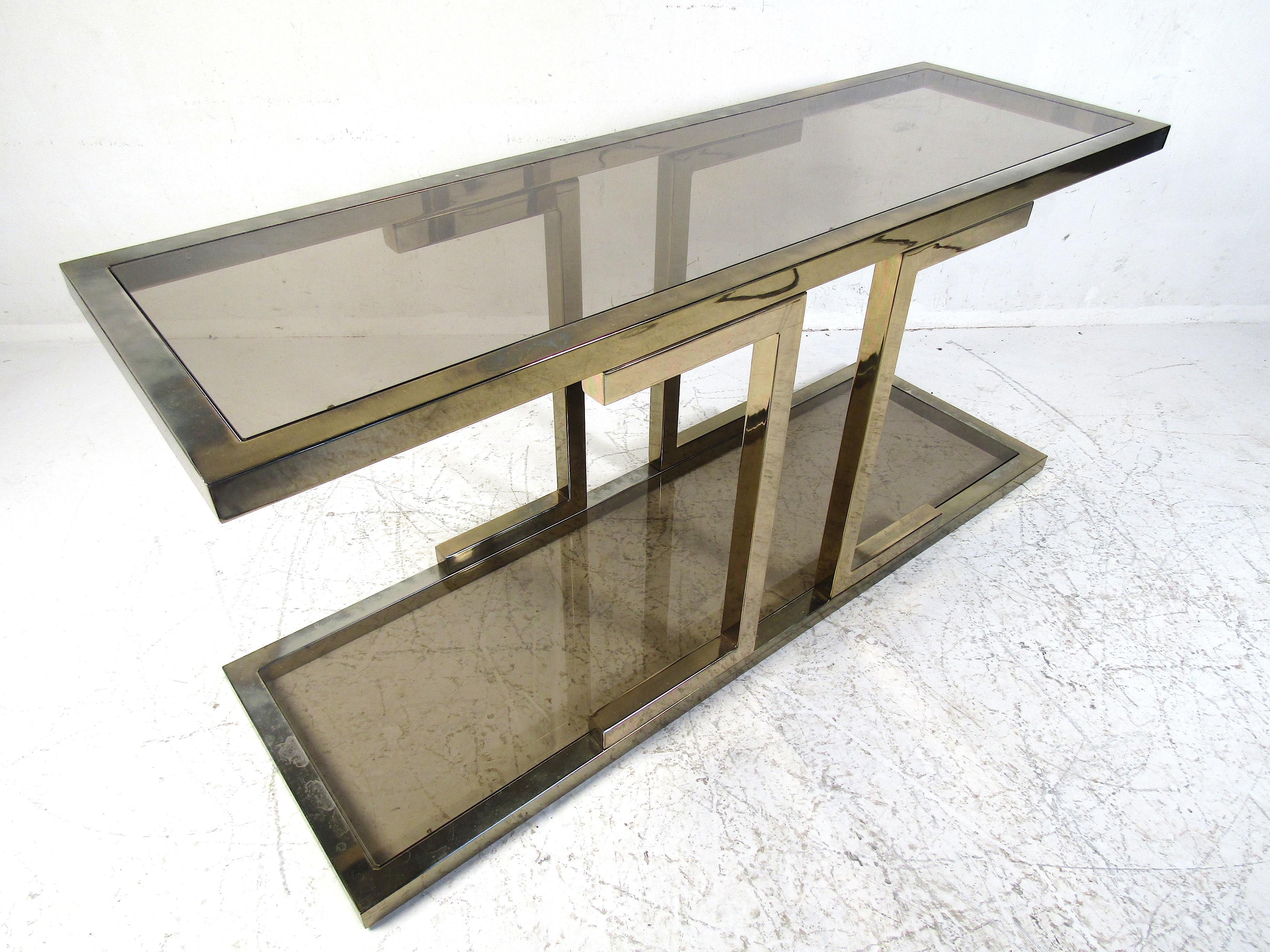 Stylish console table with a brass frame and a pair of smoked glass inserts. Two-tiered table with an interesting design. Sure to be a nice addition to any modern interior. Please confirm item location with dealer (NJ or NY).