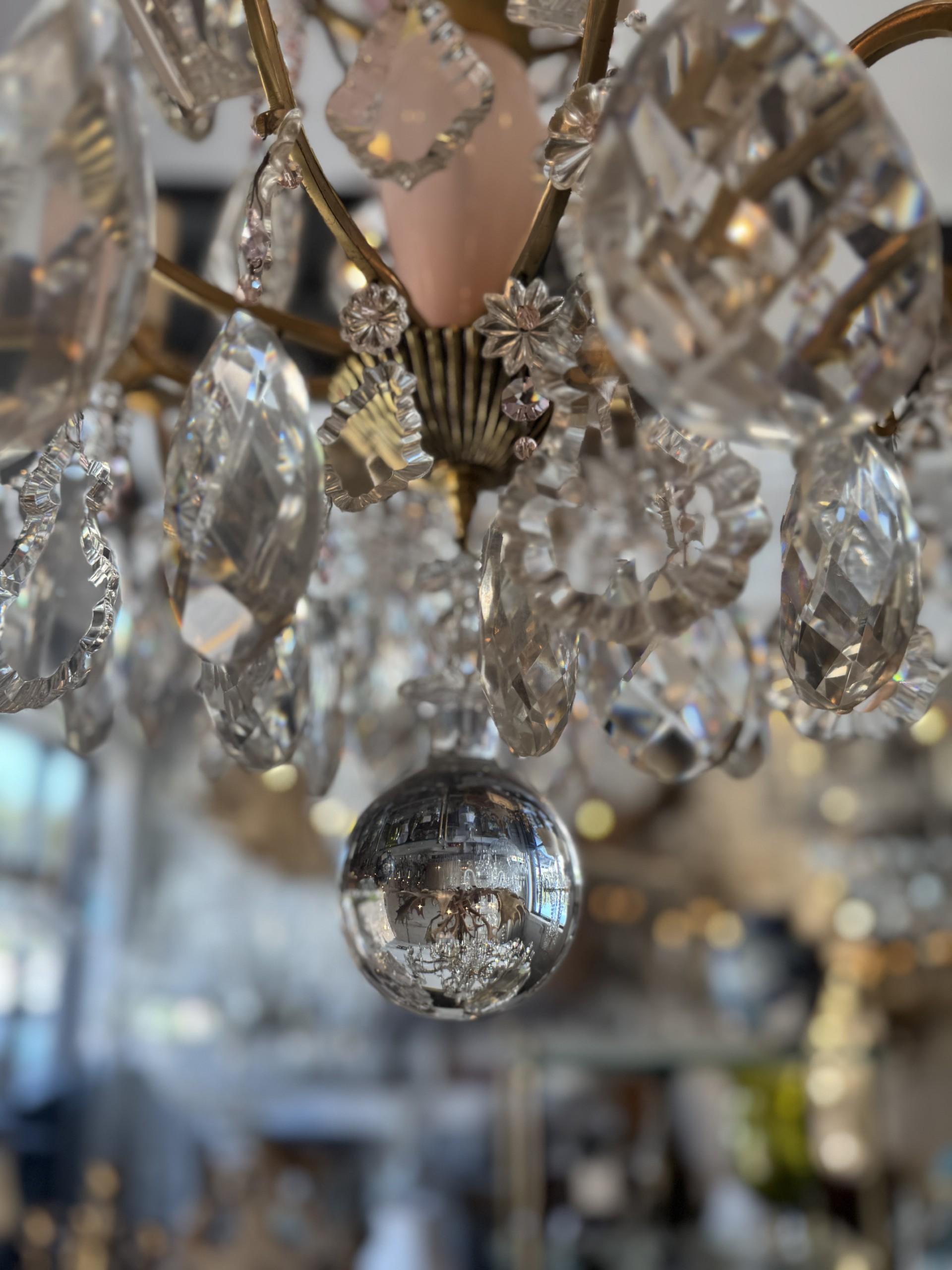 Ce magnifique lustre vintage présente des prismes de cristal étincelants, des accents floraux délicats et une colonne centrale rose tendre qui ajoute une touche de charme et d'élégance. Le cadre en laiton peut accueillir 10 lampes, créant ainsi une