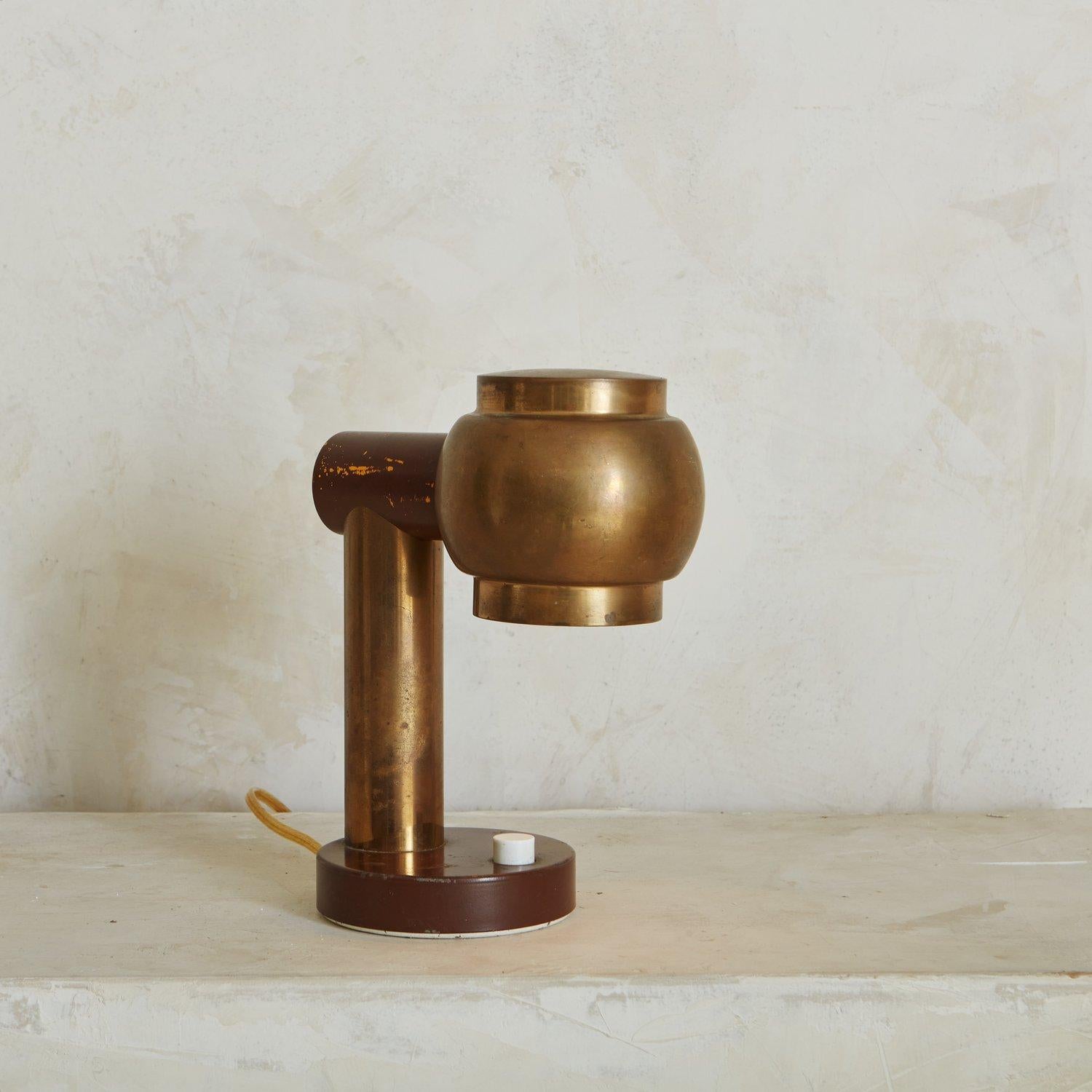 A handsome brass desk lamp featuring a thick cylindrical body and a round sculptural shade. This lamp has a round base with a white acrylic power button and is partially painted a beautiful maroon hue. We love the patina on this vintage beauty.