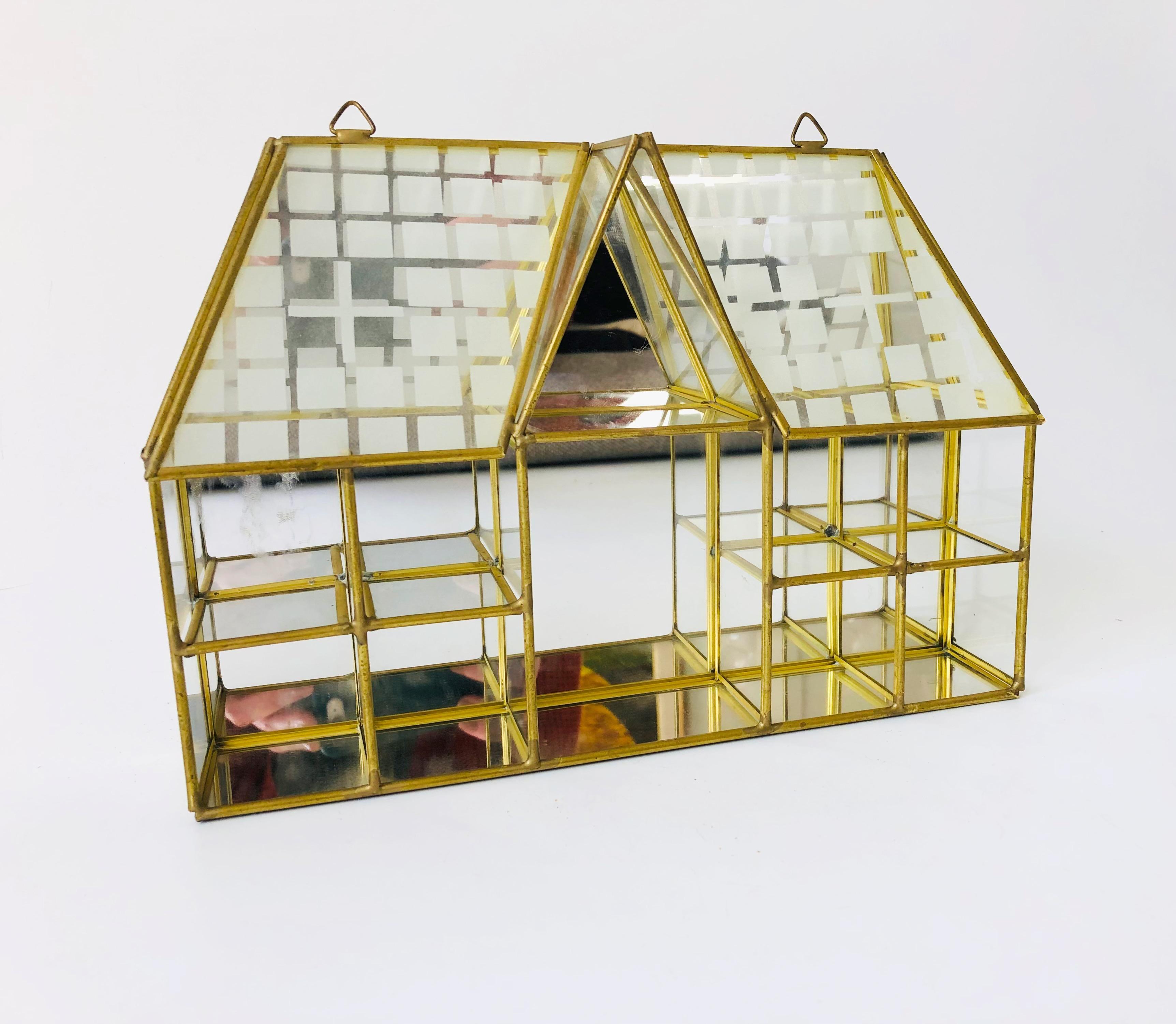 An adorable brass curio display in the shape of a house. 10 compartments of glass shelves with a mirrored back. Whimsical detailing with frosted glass shingles and windows at the top. Ready to hang by 2 hangers at the top of the shelf.
