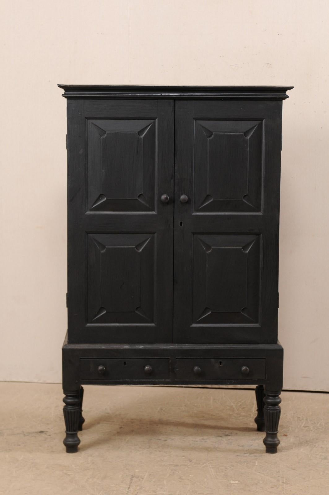 A British Colonial black colored storage cabinet from the mid-20th century. This vintage cabinet features an upper case decorated with geometrically-designed recessed panel front doors and sides, with nicely molded cornice at top. The skirt is made
