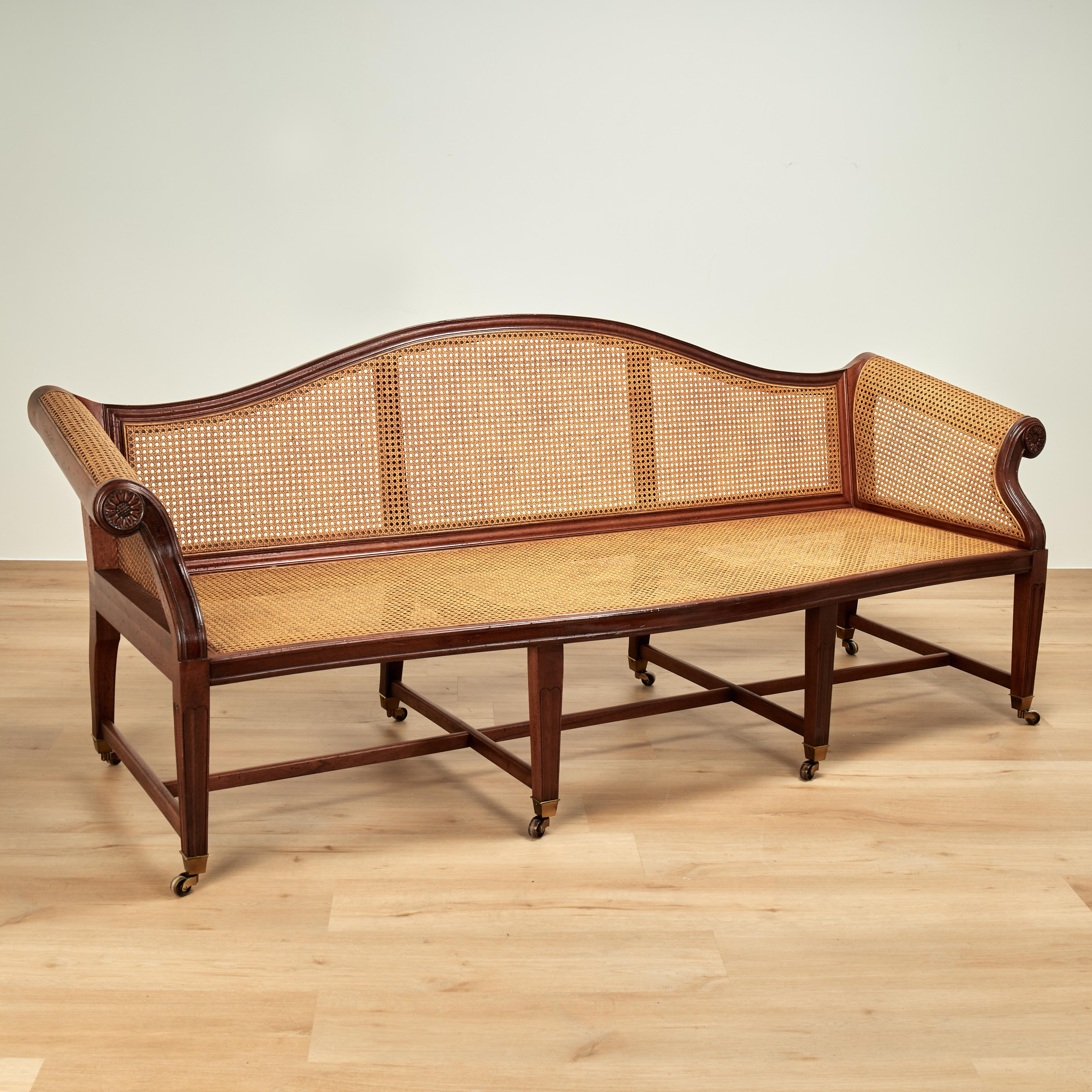 Divano in stile coloniale britannico in mogano dalle forme classiche, con schienale e seduta a canne sotto un cuscino imbottito e rimovibile, braccioli a rollo con medaglioni intagliati e gambe scanalate affusolate con rotelle in ottone.