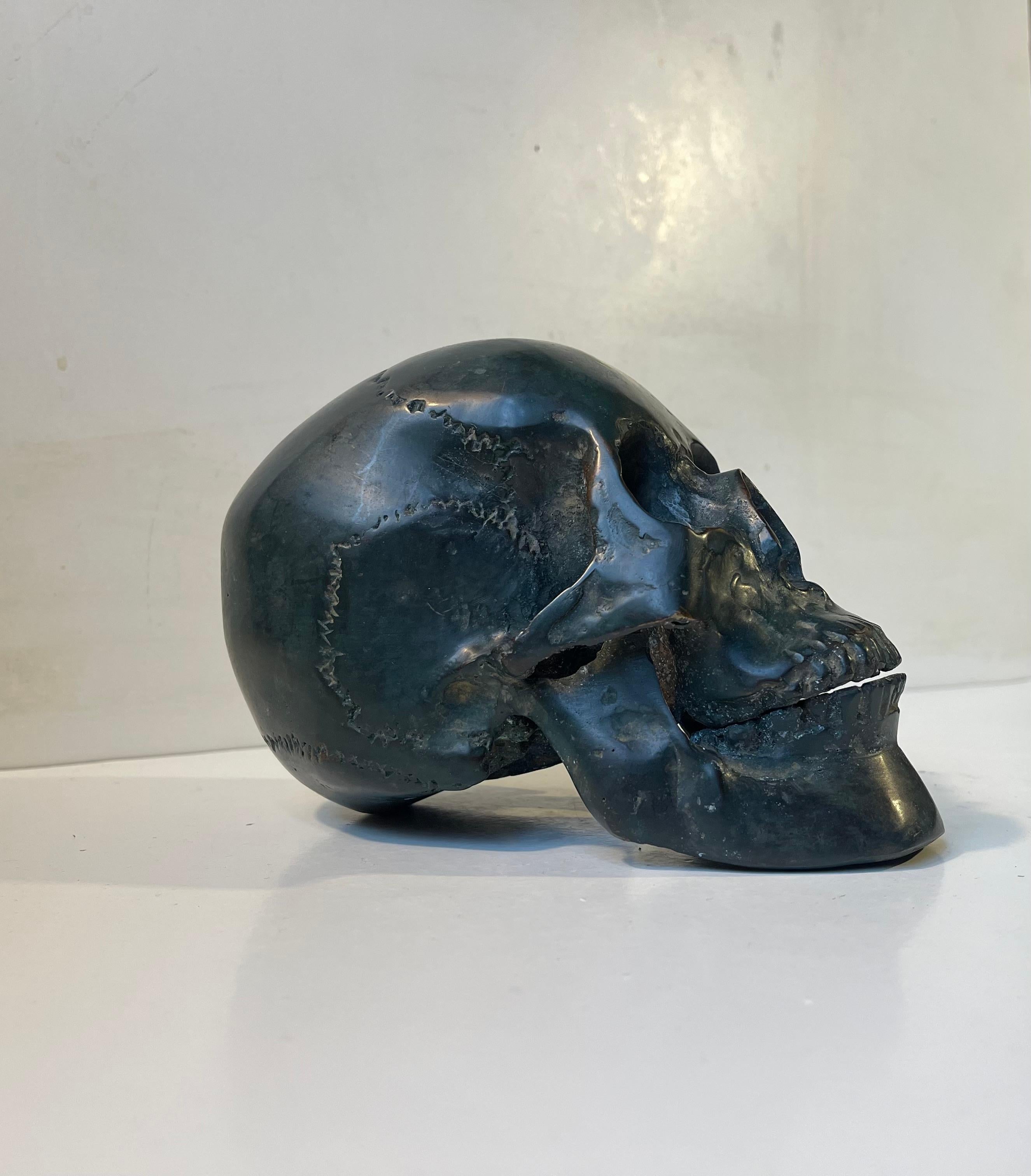 A 1:1 cast model of a human skull in patinated bronze. Its made from a cast of a real human skull, probably female, and is complete with teeth, lower- and upper jaw. This particular example shows patina and ware. The skull as a symbol serves as a