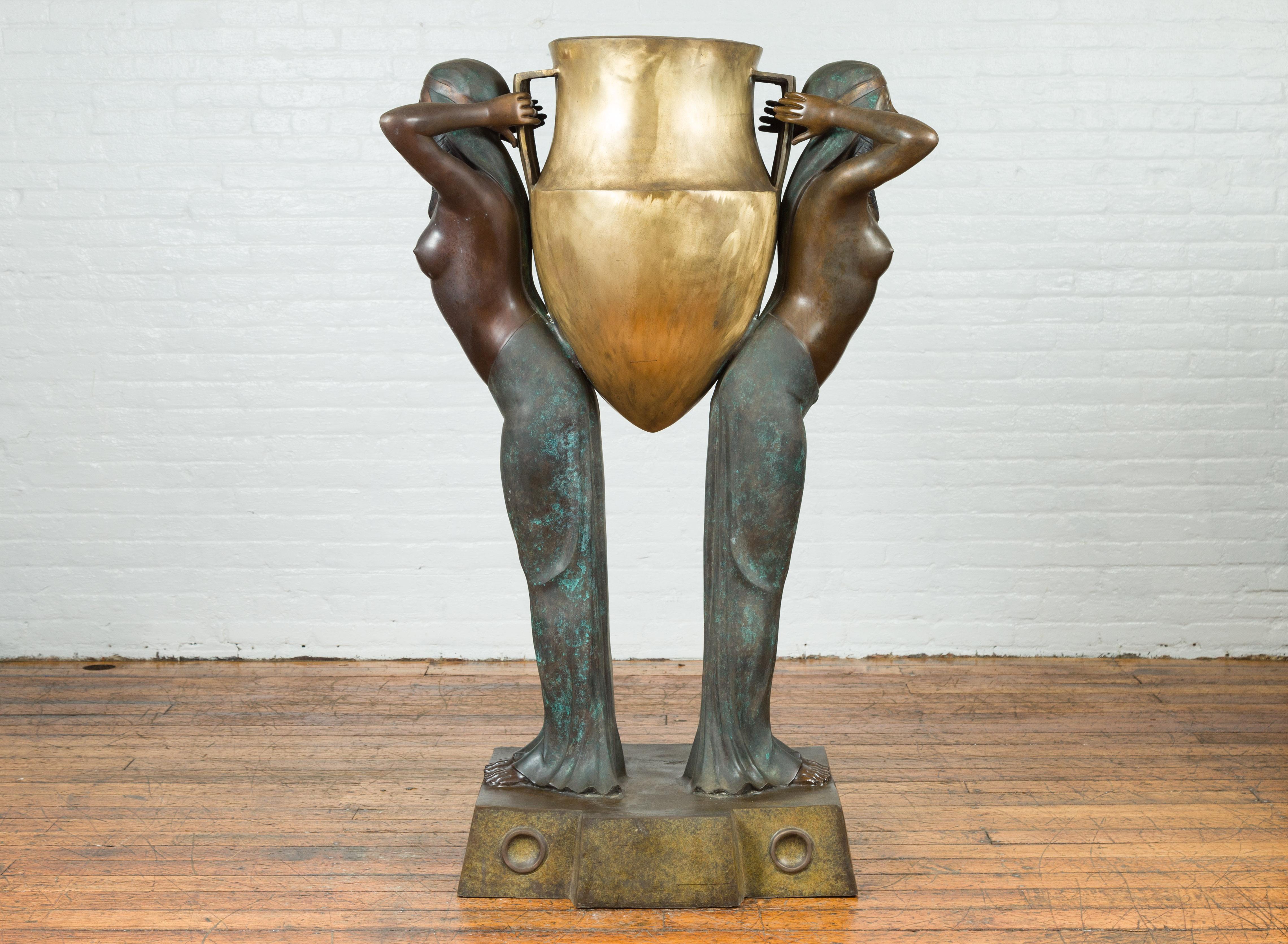 An Egyptian style vintage bronze planter depicting two bare-chested maiden carrying a double handled urn. Our eyes are immediately drawn to this striking planter depicting a scene from Egyptian antiquity. Created with the traditional technique of