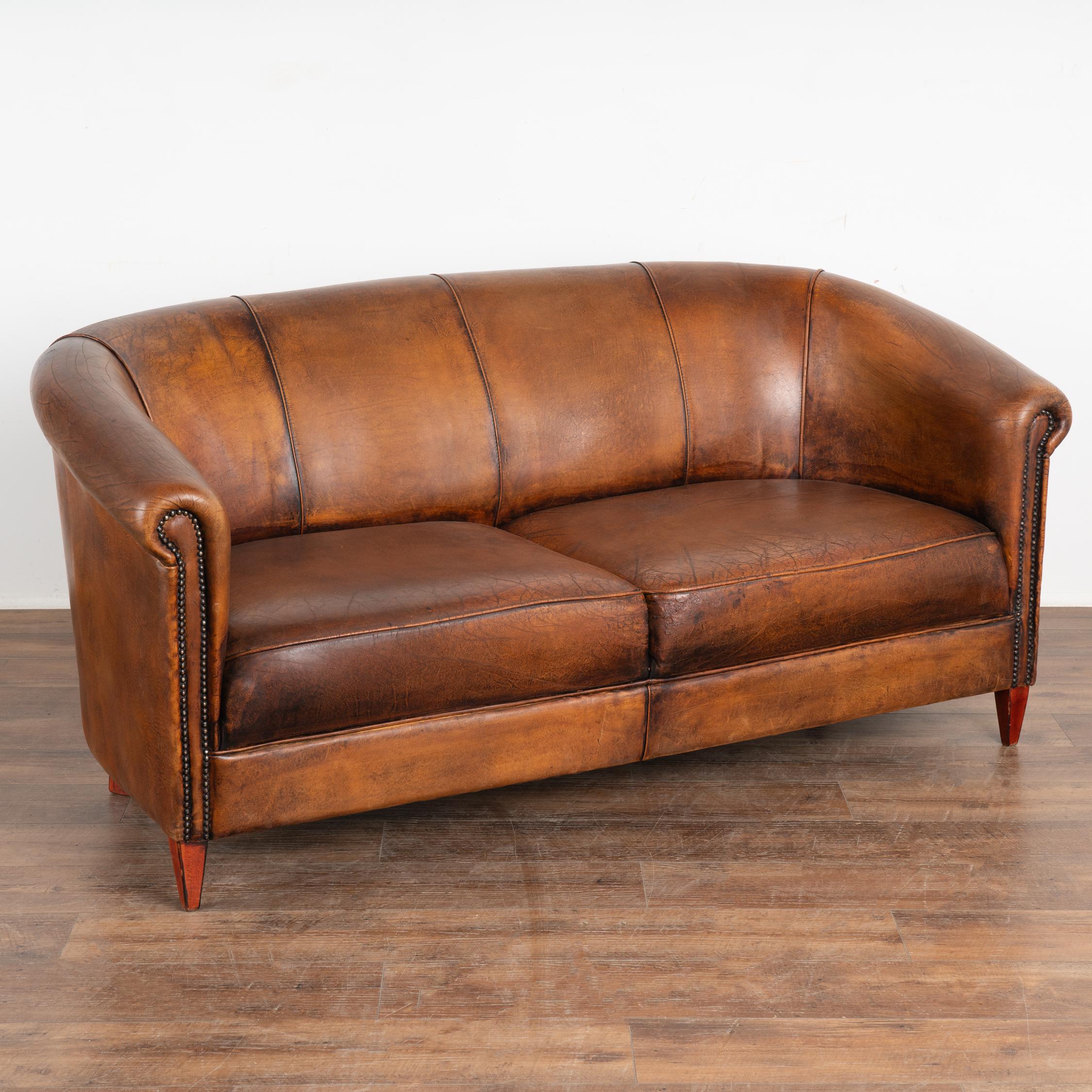 Vintage leather has an appeal all its own, especially when worn and weathered as seen in this handsome two-seat sofa. The aged patina of the brown leather has deepened through generations of use.
The gently rolled arms have traditional nailhead