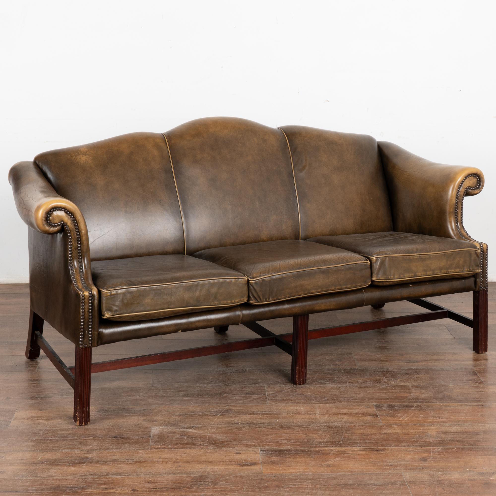 Canapé trois places en cuir marron vintage, avec auto-pipage le long du dossier et coussins d'assise détachés. Pas de marque de fabrique.
Les années d'utilisation sont révélées par la patine vieillie du cuir, y compris les impressions, les