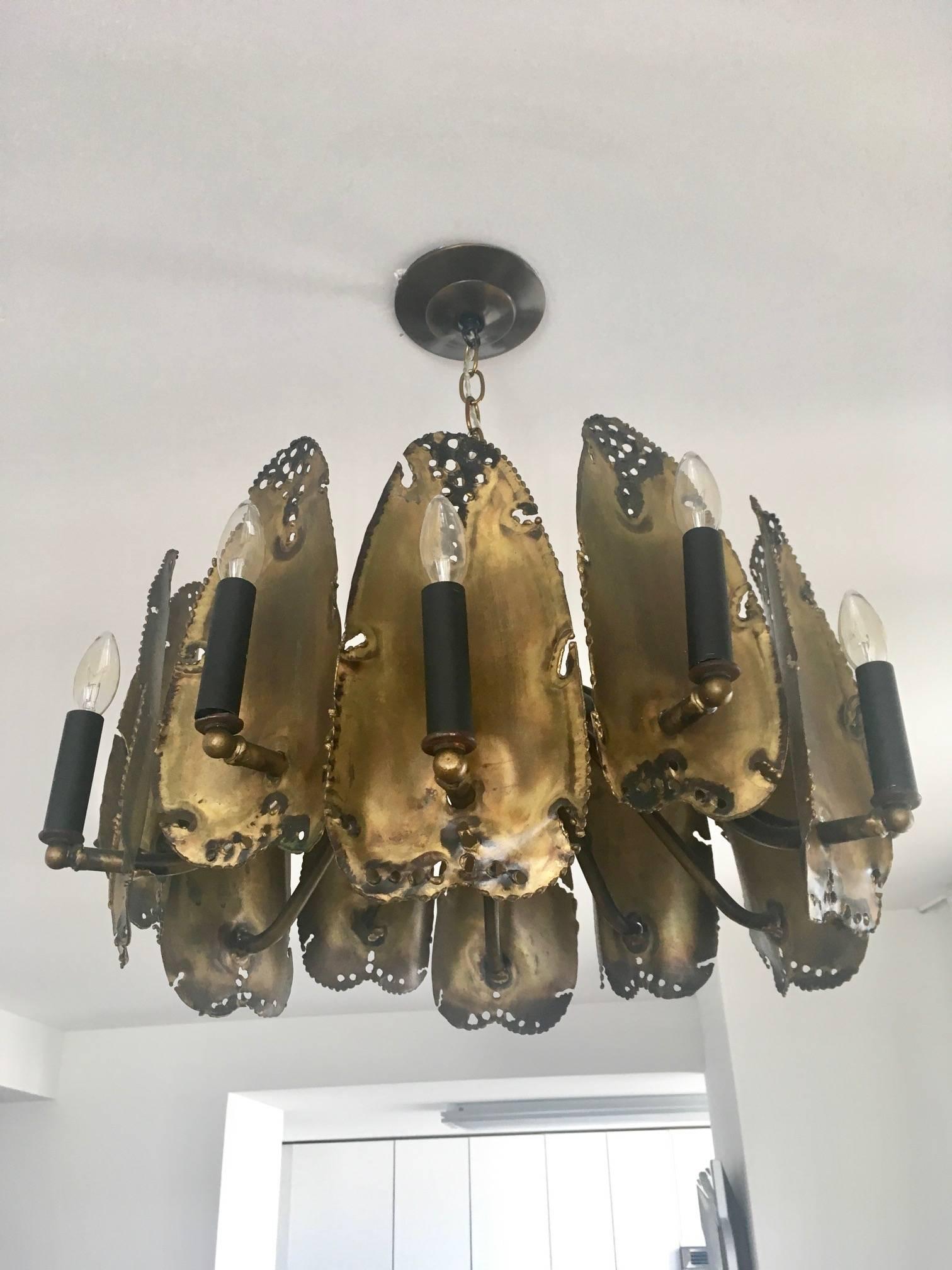 Mid-Century Modern chandelier with Brutalist design by noted metalworker and artist, Tom Greene. Made of welded brass metal and iron with a torch flame finish and with pierced details. Sculptural design fitted with 12 stylized leaves or shields with