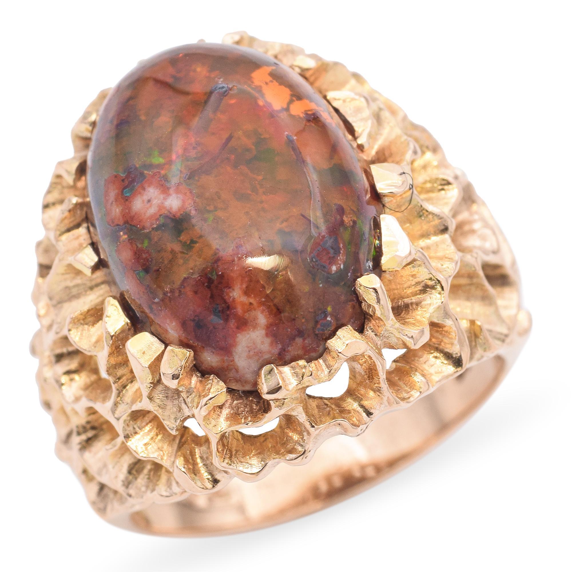 Circa 1970, this vintage ring showcases a striking fire opal cabochon, encased in an intricate textured gold setting. The opal displays a vibrant play of colors, with flashes of red, green, and orange. The gold setting features a ruffled, organic