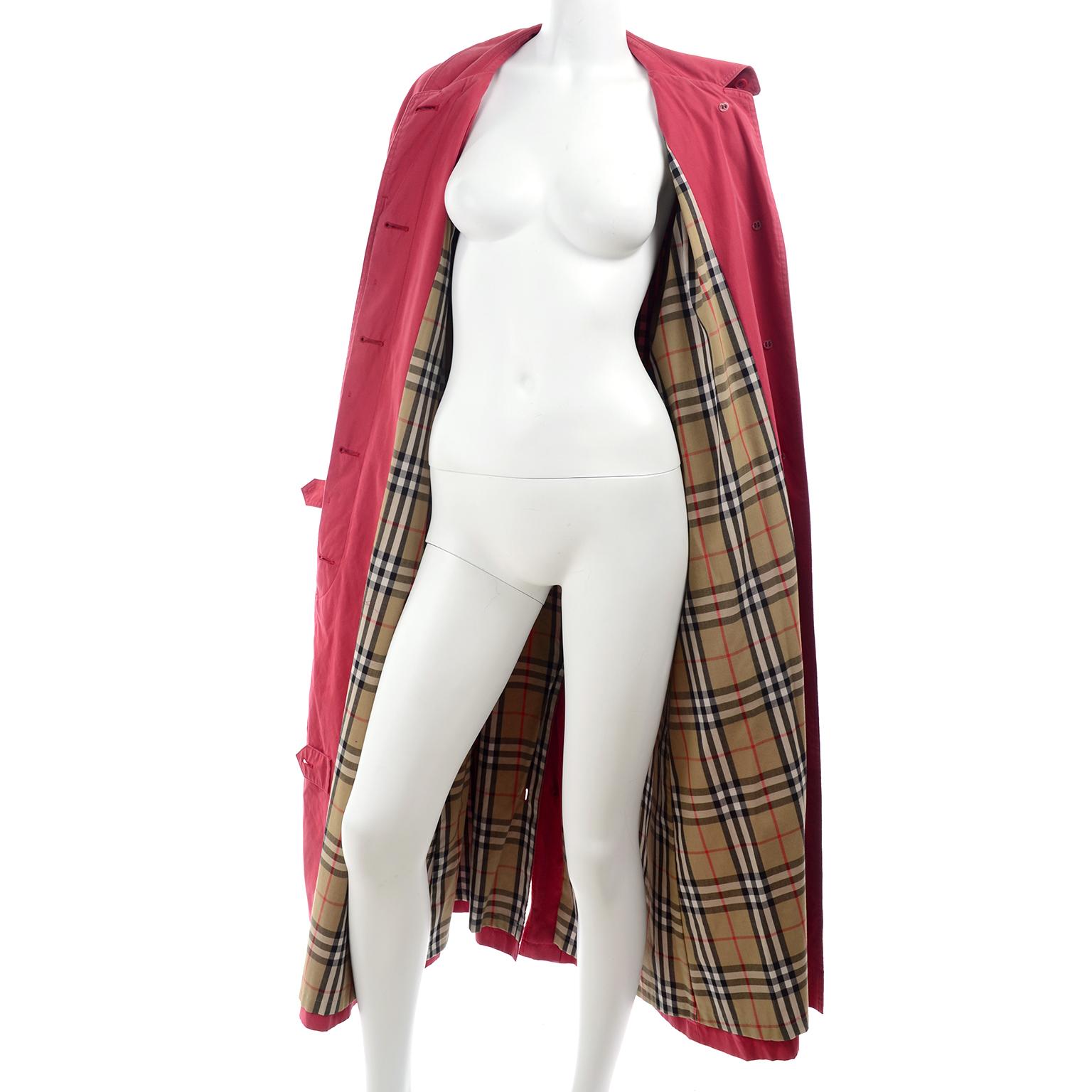 This great vintage Burberrys of London trench coat is in a raspberry pink red fabric lined with the classic red, tan, and black Haymarket Check tartan plaid Burberry print. Front button closure with a small slit in the back with a button closure as