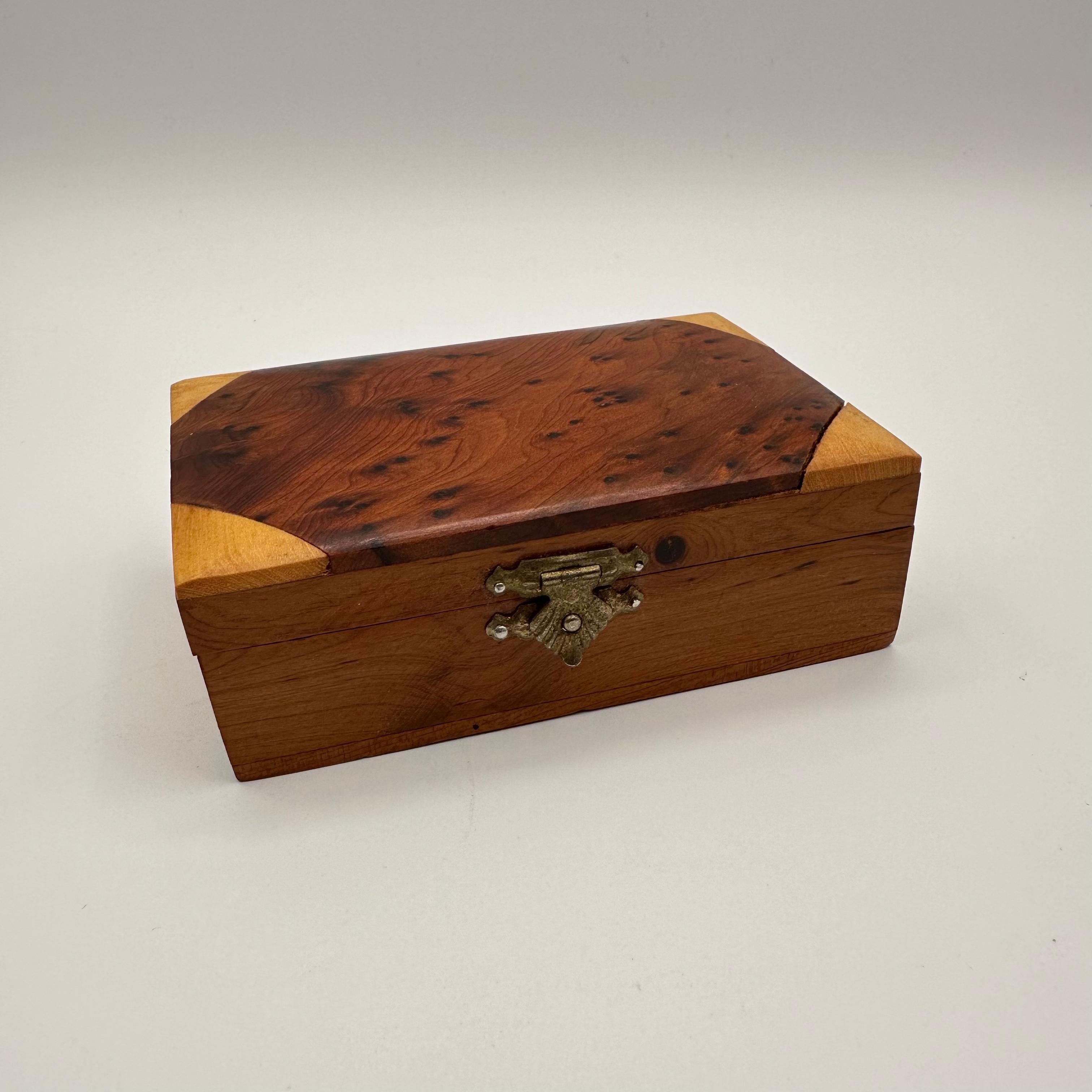A small vintage rectangular lidded box with multiple species of wood. Primarily in a darker wood with small burls, featuring inlaid triangle shaped corners in a lighter wood tone. The lid is hinged and closes with a small brass clasp with a