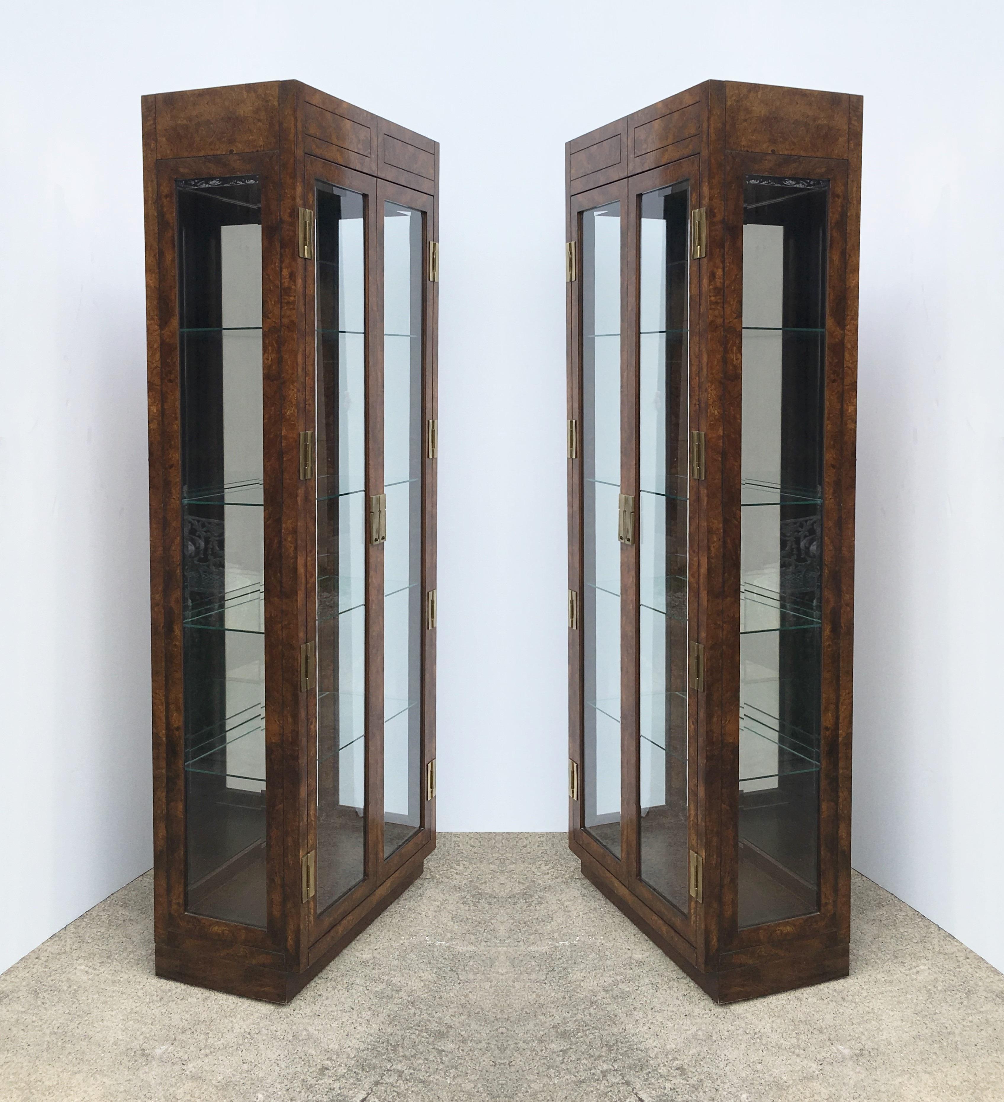 Pair of Campaign style display cabinets. The cabinets feature two beveled glass doors which open to an interior with a mirrored back. There are four thick glass shelves in each and one interior light. Heavy brass hardware adorns the front. Can be