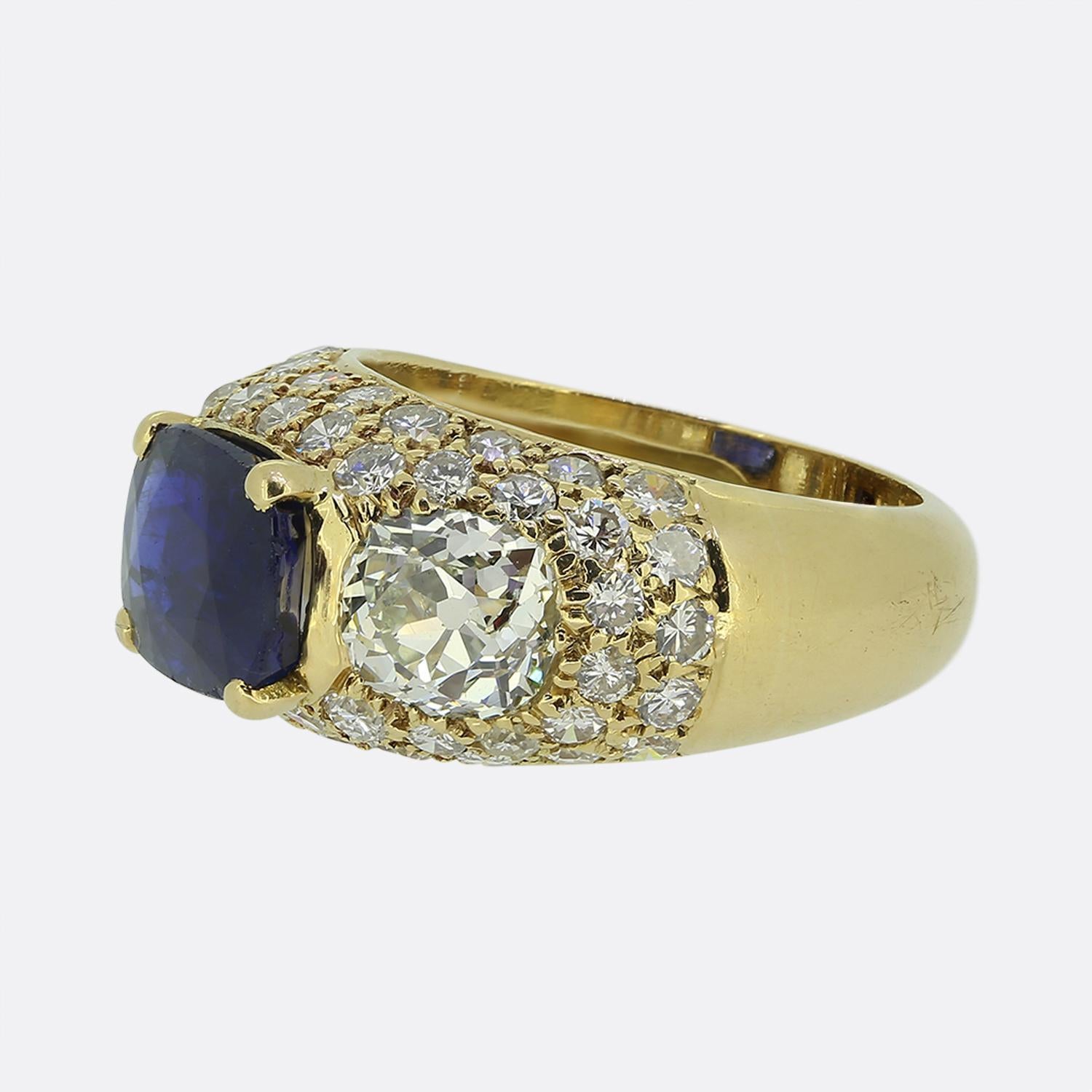 Nous avons ici une exquise bague à trois pierres en or jaune 18ct, ornée d'un saphir birman ovale à facettes au centre de la face. Ce saphir naturel d'un bleu profond est flanqué d'un duo de gros diamants taillés à l'ancienne, d'un blanc éclatant,