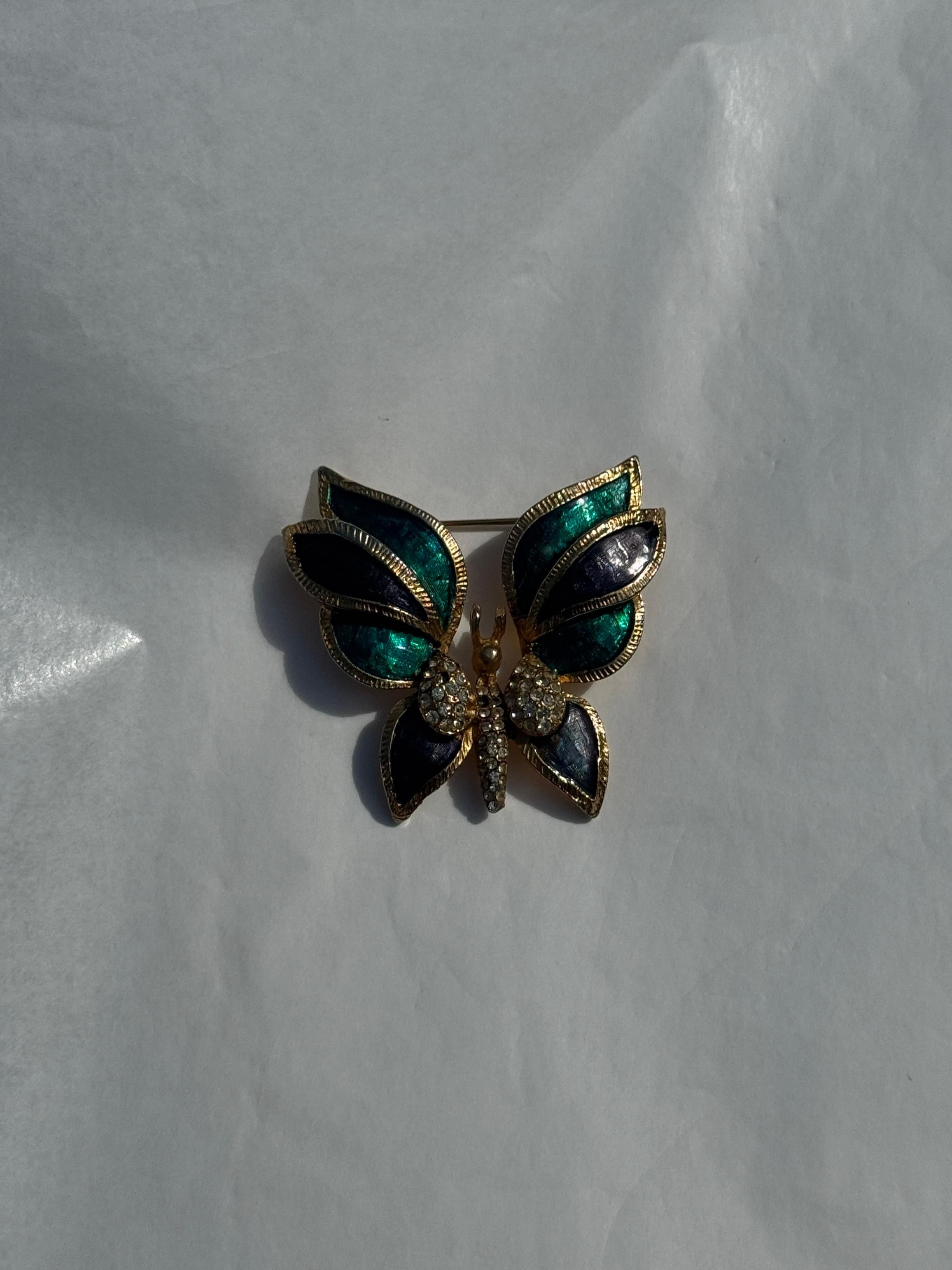 This exquisite vintage brooch, most likely from the 1960s, showcases a beautifully stylized butterfly, masterfully crafted with enamelled wings in rich shades of deep blue and emerald green. Each wing is delicately bordered in raised gold-toned