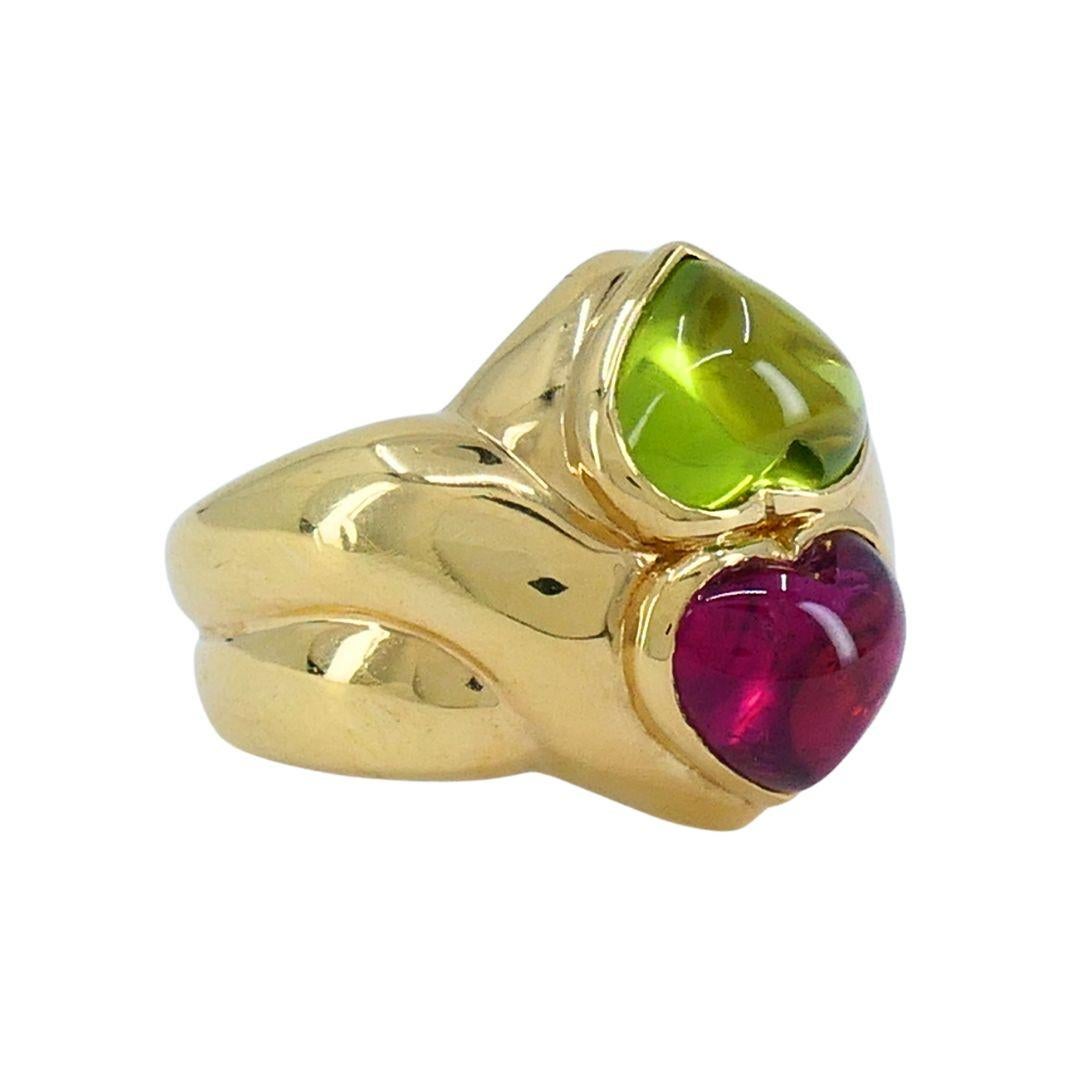 Questo anello Bulgari Cuore è realizzato in oro giallo 18 carati e presenta due gemme cabochon a forma di cuore: un peridoto verde e una tormalina rosa. Le pietre sono incastonate in un design impilato verticalmente, creando un'estetica audace e