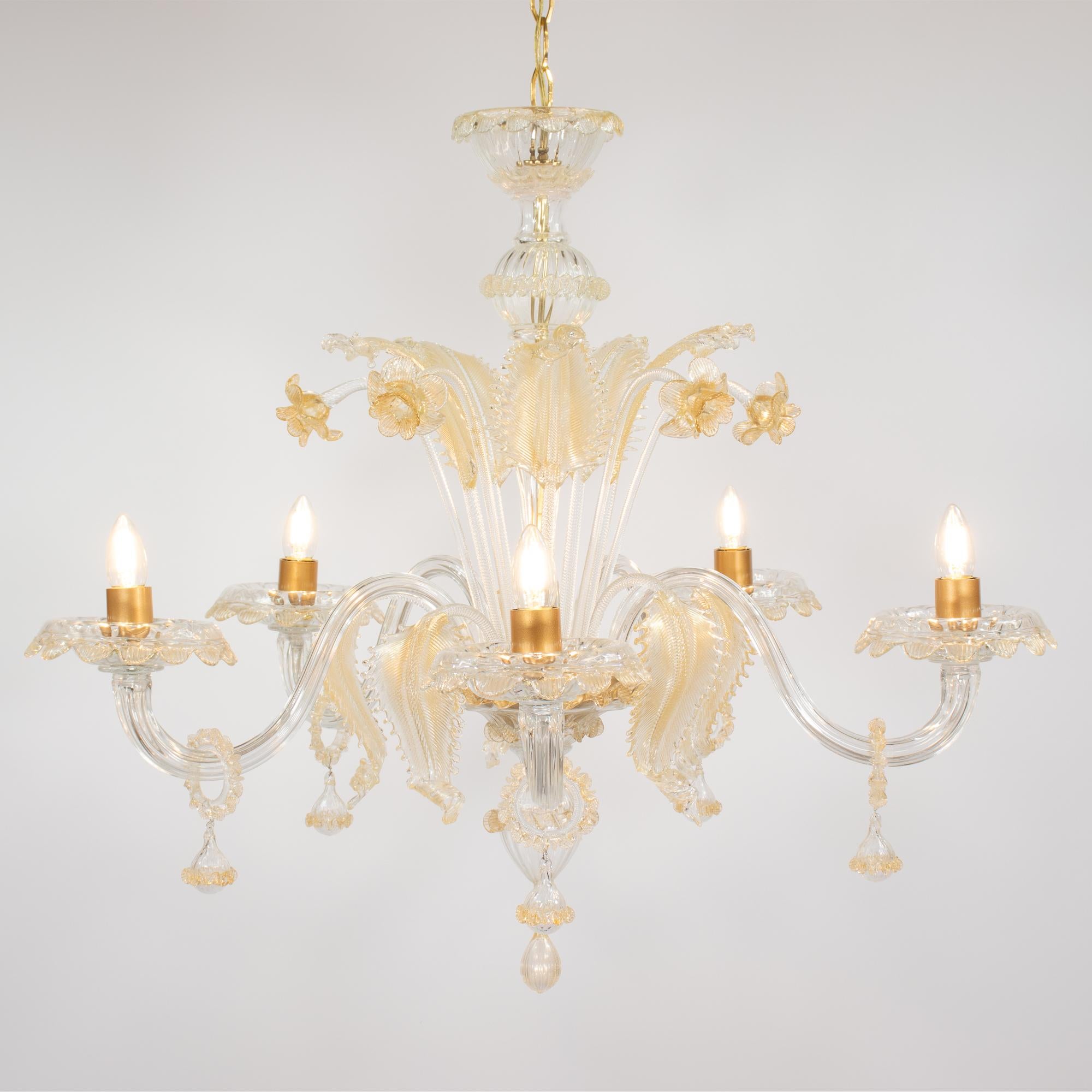 Dieser mit Gold durchzogene, mundgeblasene Cristallo Murano-Kronleuchter ist ein Beispiel für venezianische Kunstfertigkeit in Reinkultur. Das Stück verfügt über fünf anmutig geschwungene Rollenarme mit hängenden, goldverzierten Blumenkugeln, die zu