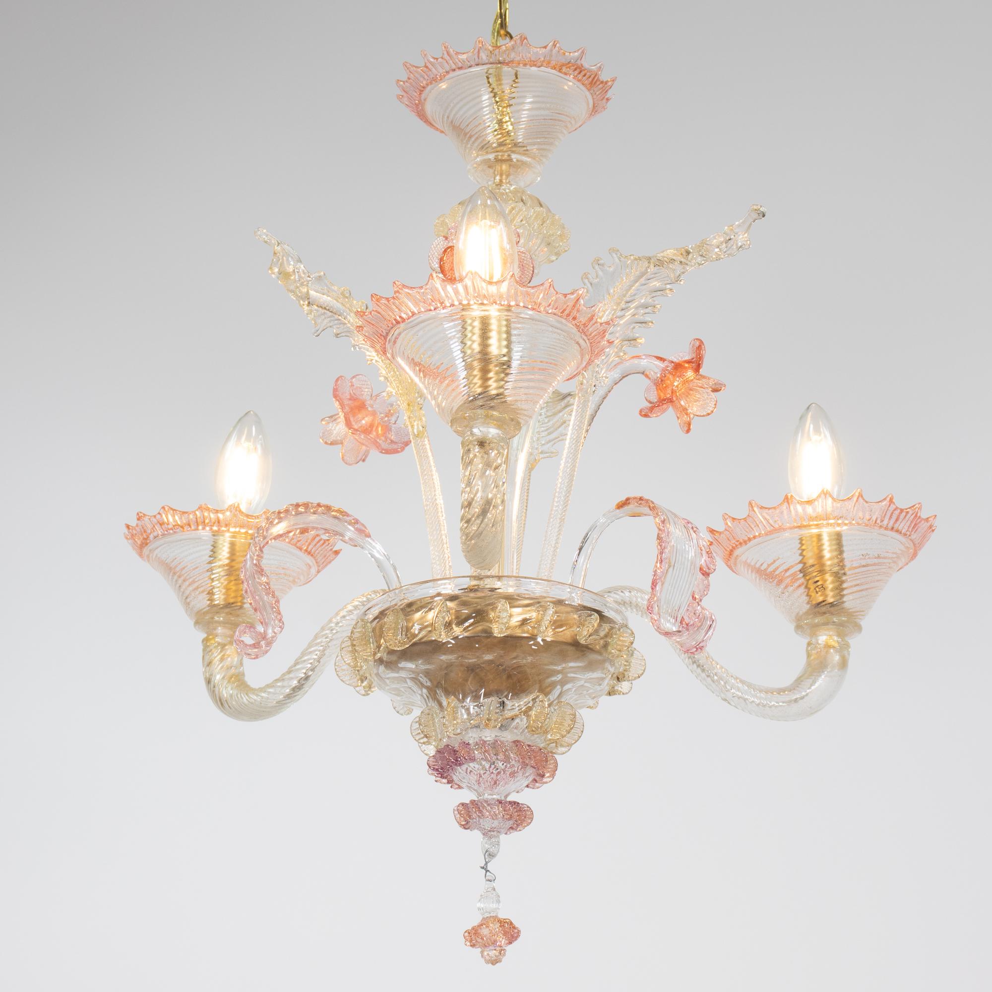 Questo lampadario in Cristallo di Murano soffiato a mano, di colore rosa e oro, rappresenta il meglio dell'arte veneziana. L'opera presenta tre bracci graziosamente ondulati che terminano in bobeches bordate di rigatino rosa, ognuna delle quali